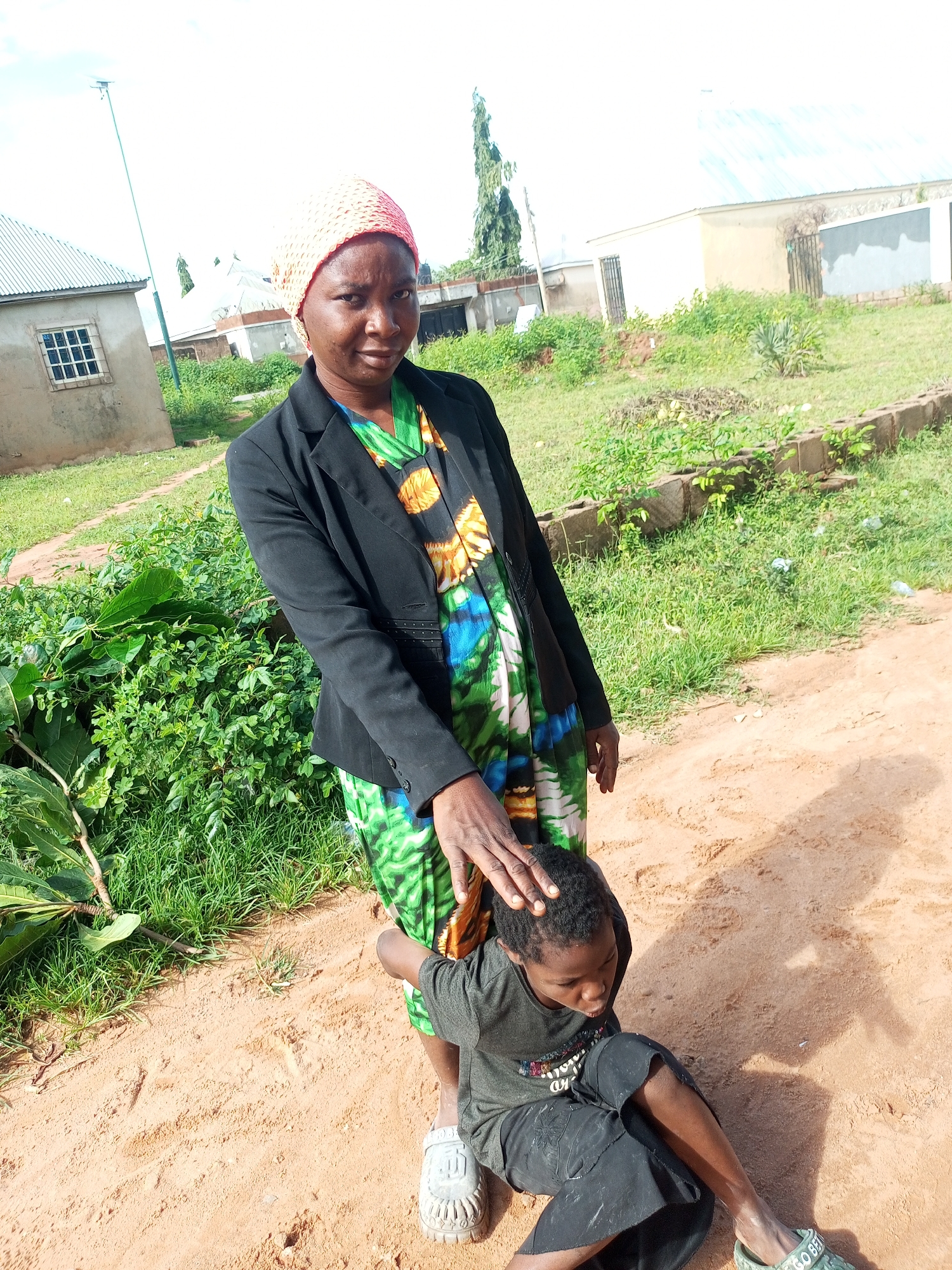 A woman stands with her hand on the head of a young child who is sitting on the ground in front of her. The woman is wearing a black blazer over a colorful, patterned dress and a light-colored headscarf. The child is wearing a dark t-shirt and dark shorts. They are outdoors in a sandy area with green vegetation and buildings in the background. The environment appears to be a rural or semi-urban setting in Tempure, Nigeria. The sky is bright and overcast.