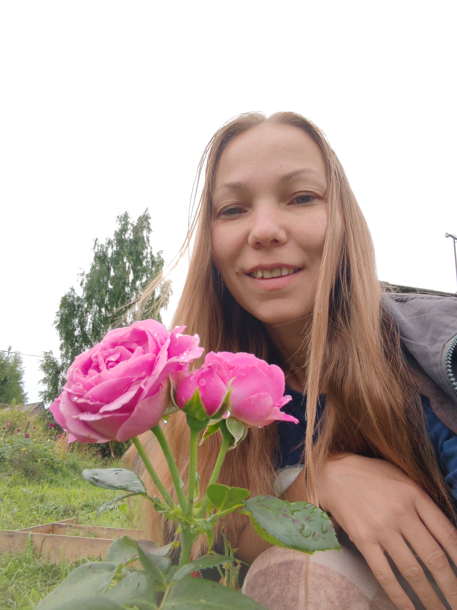 A woman with long, light brown hair is captured in a selfie, holding a pink rose with water droplets. She is smiling and looking directly at the camera. Behind her, a lush green garden with trees and plants suggests an outdoor setting. The sky is overcast. The image appears to be taken in an informal, natural environment, likely a garden or backyard. The location in Chukhlem, Russia is inferred from the provided context.