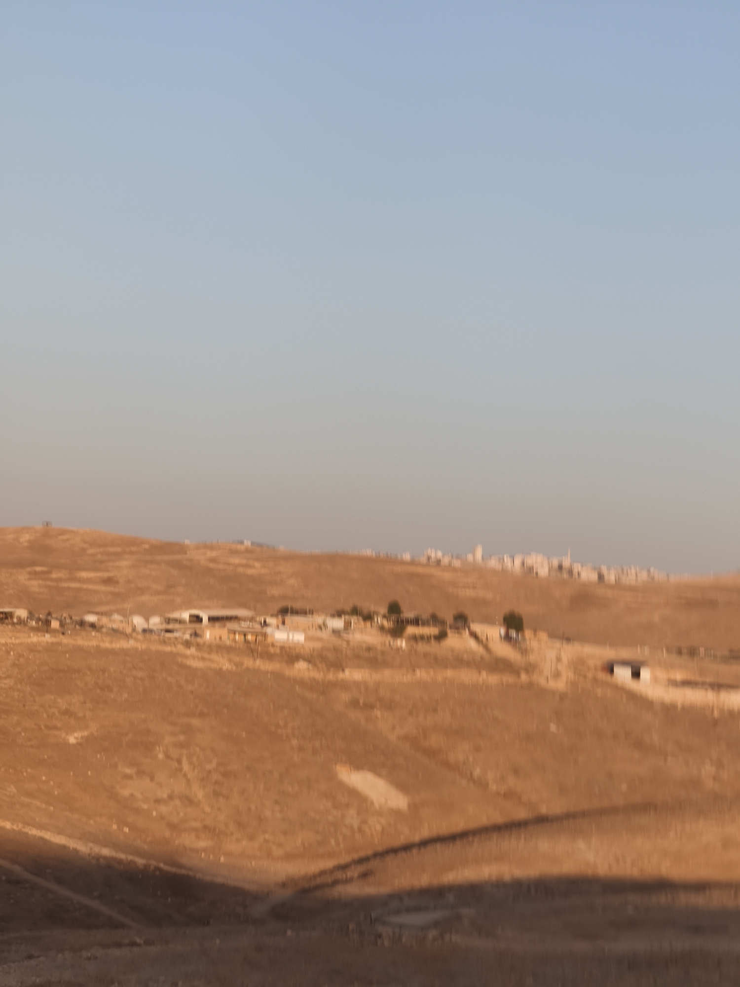The image depicts a vast, undulating landscape under a clear blue sky. The terrain is predominantly arid and appears to be dry earth and sparse vegetation, likely a hillside or valley. In the mid-ground, there are several structures visible. These include what appear to be small buildings, possibly residential or agricultural, scattered across the landscape. Some of these structures are white or light-colored, with dark roofs. Further in the distance, along the horizon, a more densely populated area with a skyline of buildings is visible, suggesting a town or city. The lighting indicates it is either early morning or late afternoon, with long shadows cast across the terrain, highlighting the contours of the land. The overall impression is one of a rural or semi-rural setting within a potentially inhabited region.