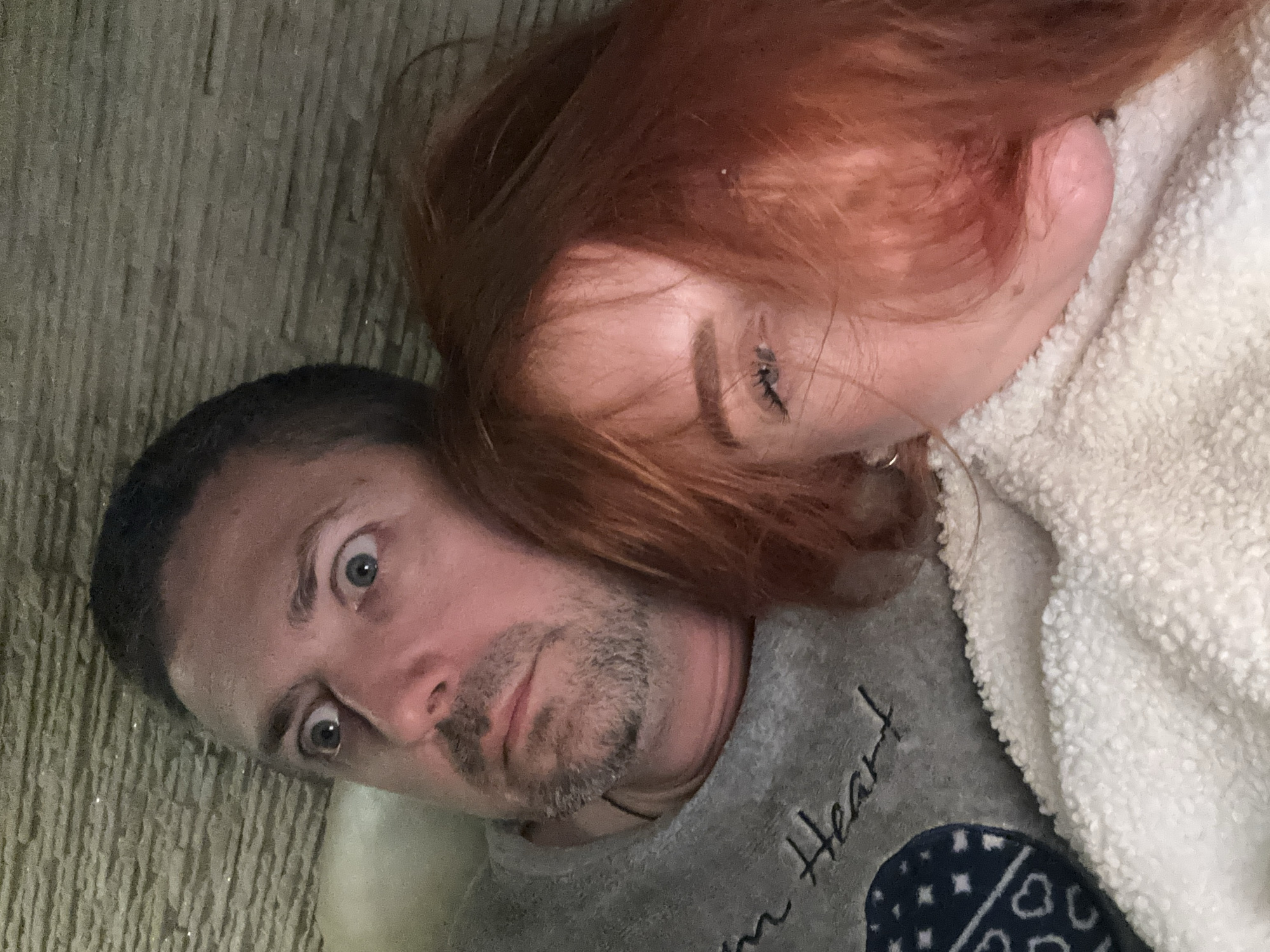 A candid, rotated portrait features two individuals, a man and a woman, in a close-up indoor setting, likely in Budva, Montenegro. The man, positioned in the foreground and appearing to lie on his back, dominates the left side of the frame. He has short dark hair and stubble, with striking light blue eyes wide open, looking directly at the camera with an expression that suggests surprise or playful alarm. He wears a grey sweatshirt with partially visible text, "m Heart," and a blue and white patterned design near the bottom right.
Resting closely beside him, mostly obscured by her flowing reddish-brown hair and a white, fluffy blanket or towel, is the woman. Only a portion of her face is visible, with one eye partially closed, showcasing long, dark eyelashes. Her posture and expression convey a sense of relaxation or sleepiness, contrasting with the man's alertness. Her ear is visible, nestled against the white fabric.
The background consists of a textured surface, appearing to be a carpet or a vertically grooved wall, in shades of grey and beige, possibly illuminated by soft, ambient light, making the time of day indeterminate. The intimate positioning of the two subjects suggests a moment of shared rest or affection.