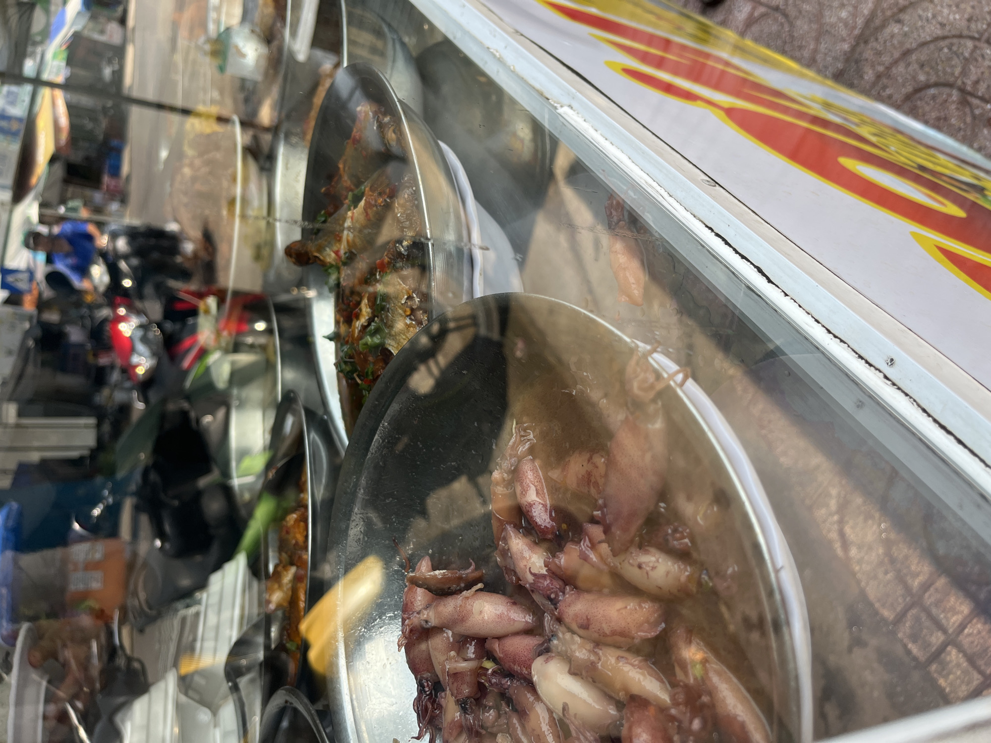 The image shows several stainless steel trays filled with various cooked seafood dishes inside a glass display case, likely part of a street food vendor stall in Bac Nha Trang Ward, Vietnam. The tray closest to the camera contains small squids. Behind this, a tray contains cooked fish. Reflections in the glass obscure the background, showing a street scene with motorcycles and parts of buildings. The top of the display case is visible, showing a sign with red and yellow coloring. The overall impression is of a typical Southeast Asian street food scene.