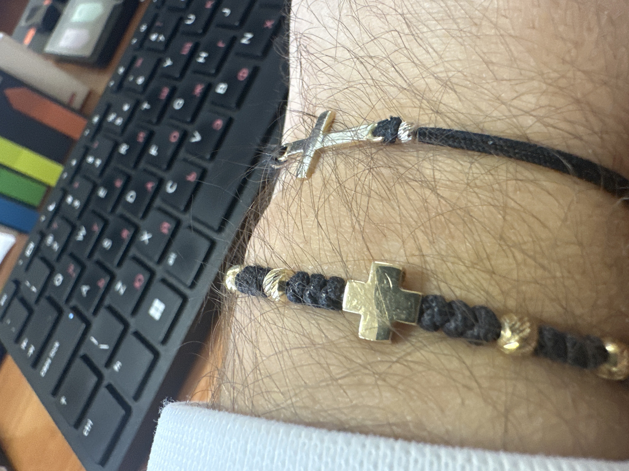 The image shows a close-up of a hairy wrist, with two black cord bracelets featuring cross pendants. One bracelet has a silver cross and smooth cord, while the other has a gold cross and beaded cord with gold accents. The background includes a black computer keyboard, and a small portion of a white shirt cuff is visible. There is no information indicating this image was taken in Plovdiv, Bulgaria.