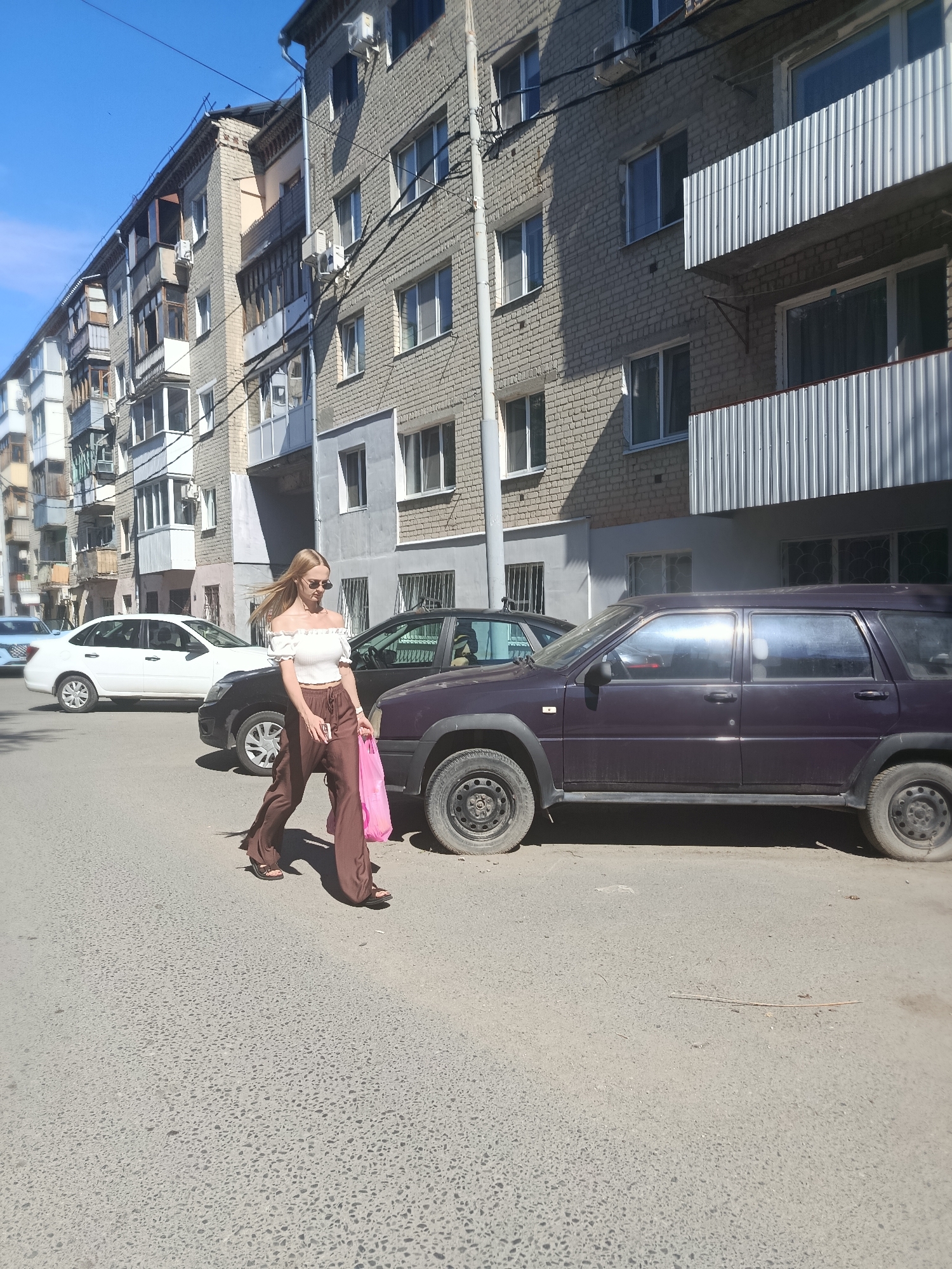 A young woman with long blonde hair is crossing a street in front of a row of apartment buildings. She is wearing a white off-the-shoulder top and brown wide-leg pants, with sandals. She is carrying a pink plastic bag in her right hand and a phone in her left hand. Two cars are parked on the street, and another car is visible further down the road. The buildings appear to be typical Soviet-era apartment blocks, common in many Russian cities. The street is paved with asphalt, and there is a sidewalk in the background. The sky is blue and clear, suggesting it is a sunny day. The scene is likely a residential area in the city of Saratov, Russia, given the architectural style of the buildings and the presence of common Russian car models.