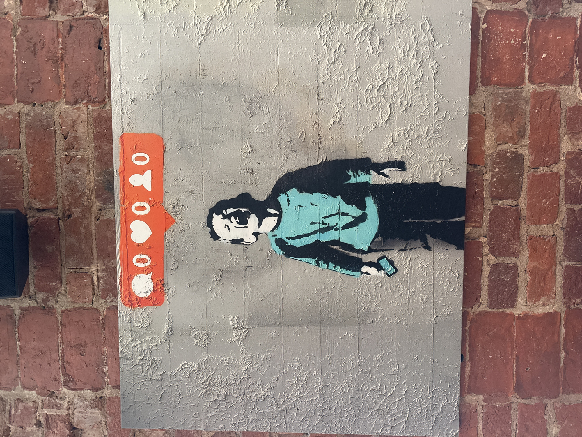 The image displays a piece of art on a brick wall, likely in Moscow, Russia, though this cannot be definitively confirmed. The artwork is a stencil depicting a young child crying, with their mouth wide open and eyes closed. The child is wearing a teal shirt and dark pants and is holding a smartphone in their right hand. Above the child's head, a red speech bubble contains social media icons: a comment bubble with "0", a heart with "0", and two silhouette figures with "0". The artwork is mounted on a grey, textured surface that resembles concrete or a painted wall. The background consists of red brickwork.