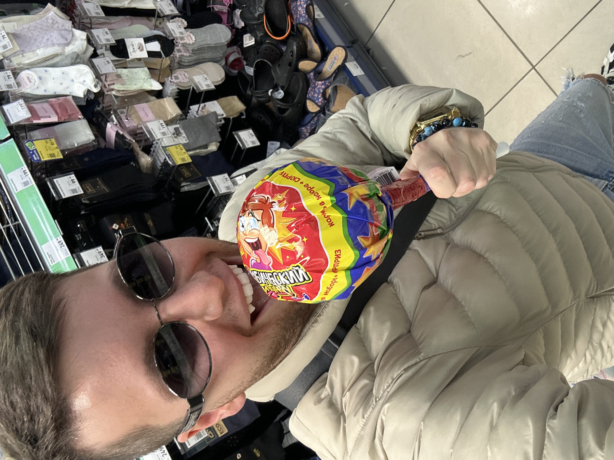 Here is a concise description of the image:
A person with sunglasses, a light-colored puffer jacket, and ripped jeans is holding a large wrapped lollipop in front of their mouth in what appears to be a store. The foreground includes shelves displaying socks and shoes. Text on the lollipop wrapper is in Russian, partially reading "в наборе + РПРИЗ" and "в наборе + СЮРПРИЗ," which indicates "in the set + prize" and "in the set + surprise." A possible location might be Russia as suggested by the Russian writing on the lollipop wrapper. The scene shows a shopping environment, suggesting a trip to a store to buy these items.