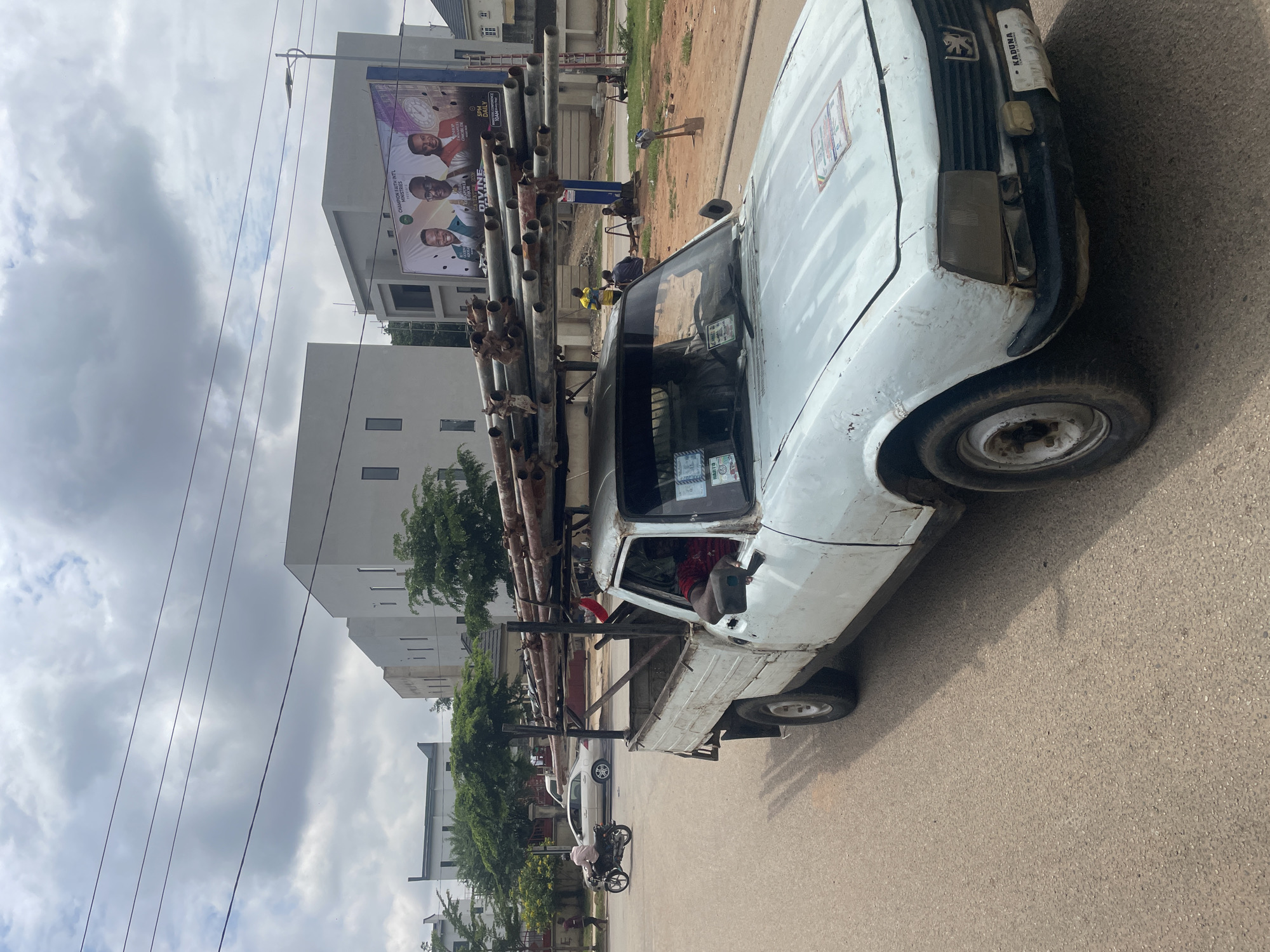 In Guzape, Nigeria, a white Peugeot pickup truck is driving on a paved road, loaded with a large quantity of metal pipes on its roof rack. The driver, a man wearing a red shirt, is visible through the driver's side window. In the background, several modern, multi-story white buildings with dark windows are visible. A billboard displaying three men and text is mounted on one of the buildings. A person on a motorcycle is seen riding away from the truck on the left side of the road, and other individuals are gathered near the roadside in the mid-ground. The sky is overcast with clouds.