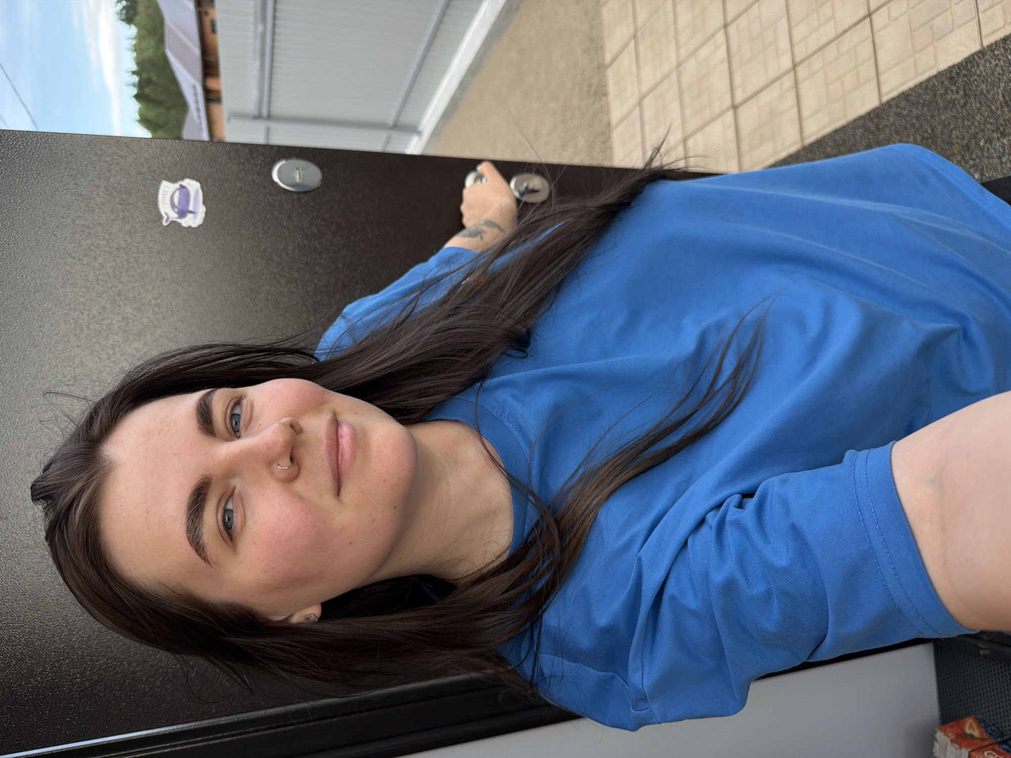 A woman with dark hair, fair skin, and blue eyes wearing a blue shirt is seen close-up in the photograph. She has a nose ring and dark eyebrows. She's positioned next to a dark-colored, possibly metal, door that appears to be an exterior door. A sticker and a round, metallic object, possibly a lock mechanism, are visible on the door. In the background, there is a glimpse of what seems to be a building with a corrugated metal roof, greenery, and what might be sky visible above the building. The location is Chaikovsky, Russia. A person's hand is visible with a tattoo on it near the door handle. The woman appears to be taking a selfie.
