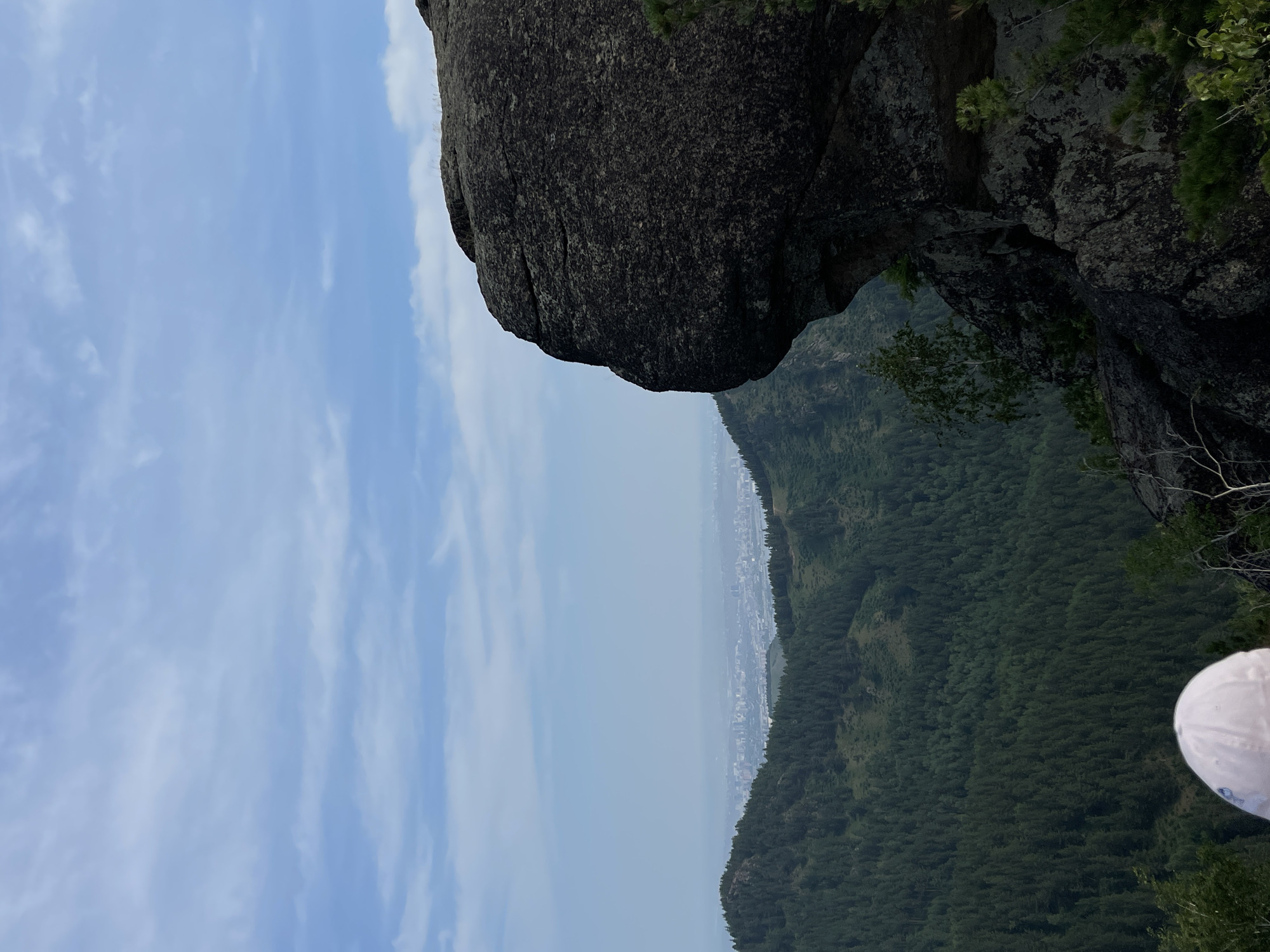 This image captures a stunning natural landscape from a high vantage point. The foreground is dominated by a large, dark, rough-textured boulder, possibly part of a rock formation or cliff. This boulder juts out over a vast, densely forested mountainside that stretches into the distance. The trees are a deep green, creating a textured carpet across the undulating terrain.
Beyond the forested slopes, a hazy urban landscape can be seen in the far distance, suggesting the presence of a city nestled within the valley or along the coast. The sky above is a clear, pale blue, adorned with wispy white clouds that streak across the frame. The overall impression is one of a serene and expansive natural vista, possibly from a hiking or scenic overlook.
The image appears to be taken during the daytime, with the natural light illuminating the scene. The orientation of the photograph is unusual, as the image is rotated 90 degrees counter-clockwise, making the sky appear on the left and the ground on the right. In the lower right corner, a portion of a white object, possibly a hat or cap, is visible, indicating the presence of at least one person observing the view. The atmosphere seems calm and peaceful, evoking a sense of awe at the scale of the landscape. No text is visible in the image.