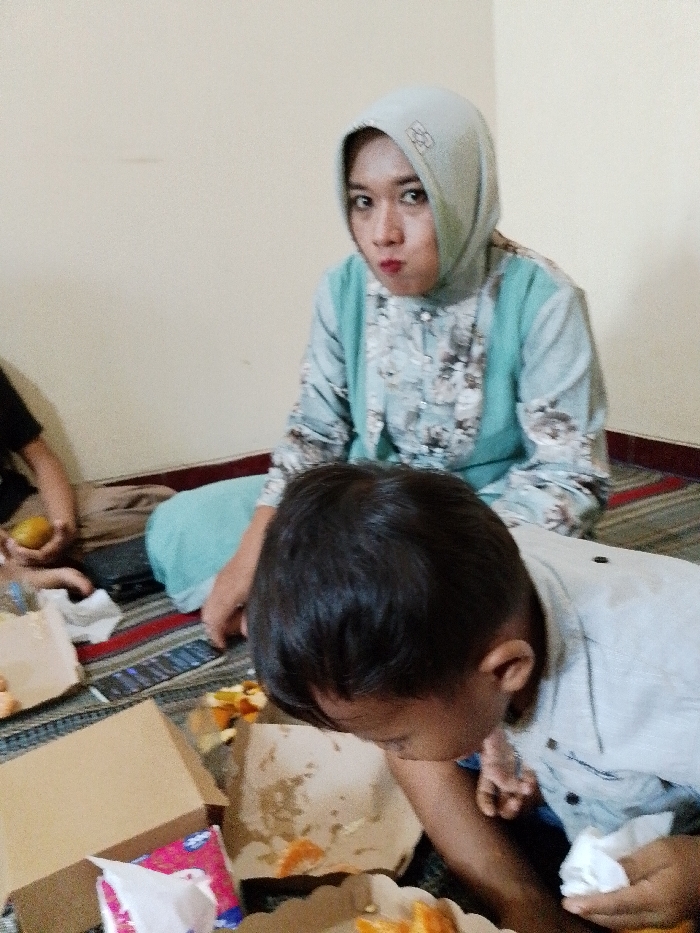 An indoor scene in Tumpangkrasak, Indonesia, depicts three individuals gathered on a striped floor mat against a light-colored wall. In the background, an adult female with a light blue hijab featuring a subtle pattern, and a patterned light blue shirt, sits facing forward with pursed lips and puffed cheeks, looking directly at the viewer. In the foreground, a young male child with dark hair, wearing a light-colored shirt, leans forward, focusing on food items. He holds a white crumpled napkin in his right hand. To the left, a third individual's arm and hand are visible, holding a yellow fruit, with part of a dark garment and light brown pants also visible. On the floor are multiple open light brown cardboard food containers, one containing a blue and white package (likely tissues or wipes), and scattered food remnants, including orange-colored pieces. A dark-colored smartphone with an illuminated screen lies on the mat near the center. A dark purse or bag is visible near the left individual's legs.