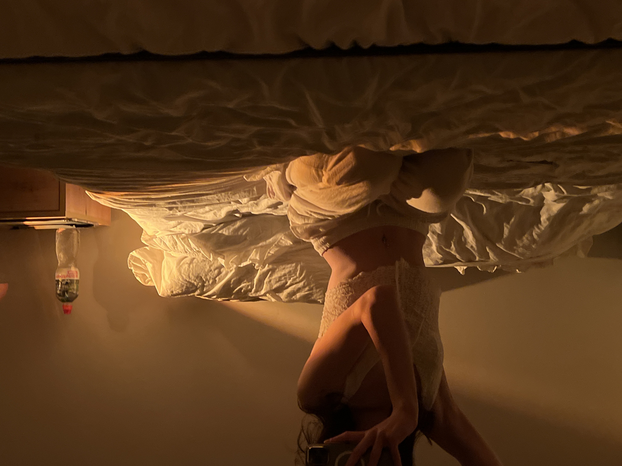 The image depicts a dimly lit bedroom scene. A person, partially visible from the chest down, is sitting on a bed covered with white and yellow linens. The person is wearing a white lace top and light-colored pants. The lighting suggests a warm, possibly evening or early morning atmosphere, with a light source casting shadows on the wall behind them. To the right of the bed, a wooden nightstand is visible, with a bottle of water and some papers on it. The overall impression is one of a private, domestic space. There is no discernible information in the image to suggest a specific location in Ternopil, Ukraine.