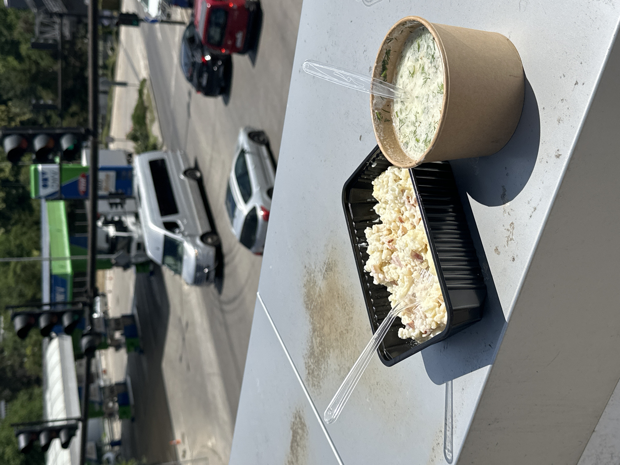 The image depicts a street scene in Veliko Tarnovo, Bulgaria, with two takeout food containers, a black rectangular plastic container filled with a creamy pasta salad and a brown paper cup filled with a soup topped with dill, placed on a grey, textured surface in the foreground. A clear plastic fork is present in the pasta salad, and a clear plastic spoon is in the soup. In the background, a multi-lane road is visible with several vehicles including a white van, a black car, a red car, and a silver car. Traffic lights are positioned above the road, and a petrol station with green and blue signage is also present. The environment appears to be outdoors on a sunny day.