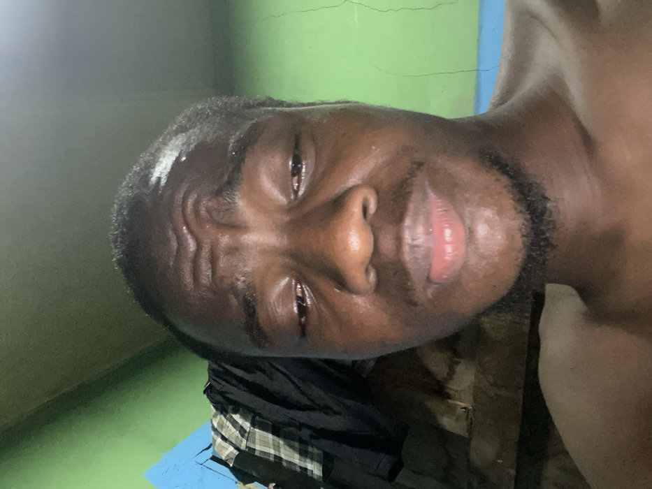 A close-up, slightly angled shot of a Black man with short hair and a beard. His forehead is wrinkled, and his skin appears sweaty or oily. He is lying on what seems to be a blue surface with a green wall behind him. Part of his upper body is visible, suggesting he might be shirtless. A black shirt and a plaid pattern are partially visible below him. The overall impression is intimate and personal.