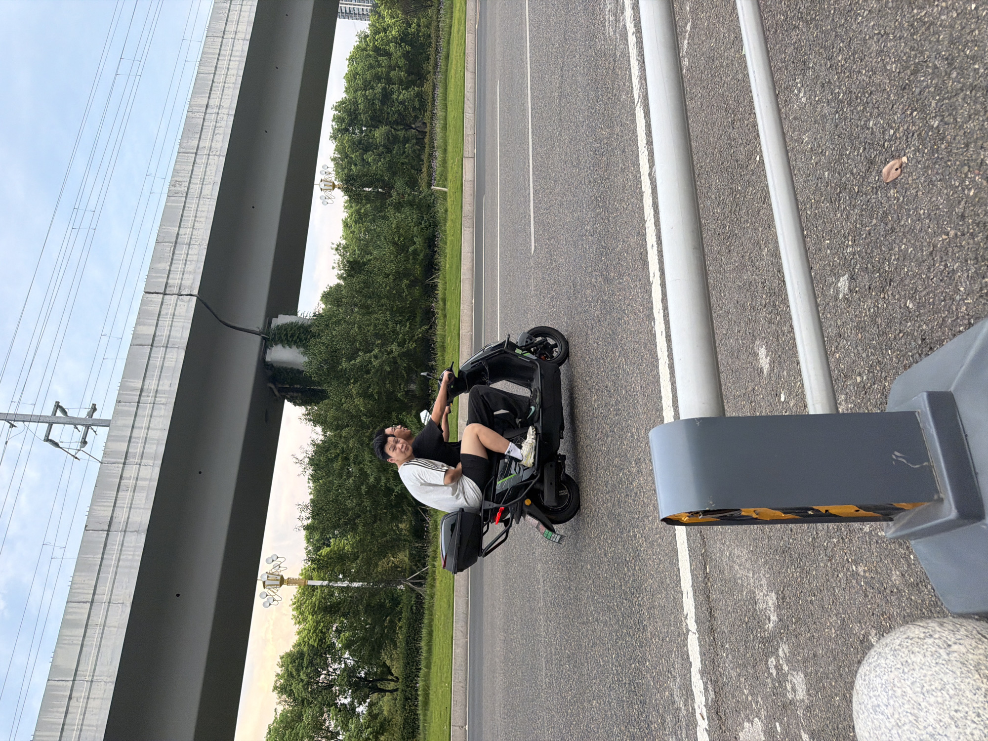 Two men ride on a black electric scooter on a road in Jiangjin, China. The man in front is wearing a white t-shirt with black stripes on the sleeves and black shorts. The man in the back is wearing a black t-shirt and black pants. The scooter has a black storage box on the back. They are riding on a paved road with white lane markings. In the background, there is a large overpass, trees, and a lamppost. The sky is cloudy.