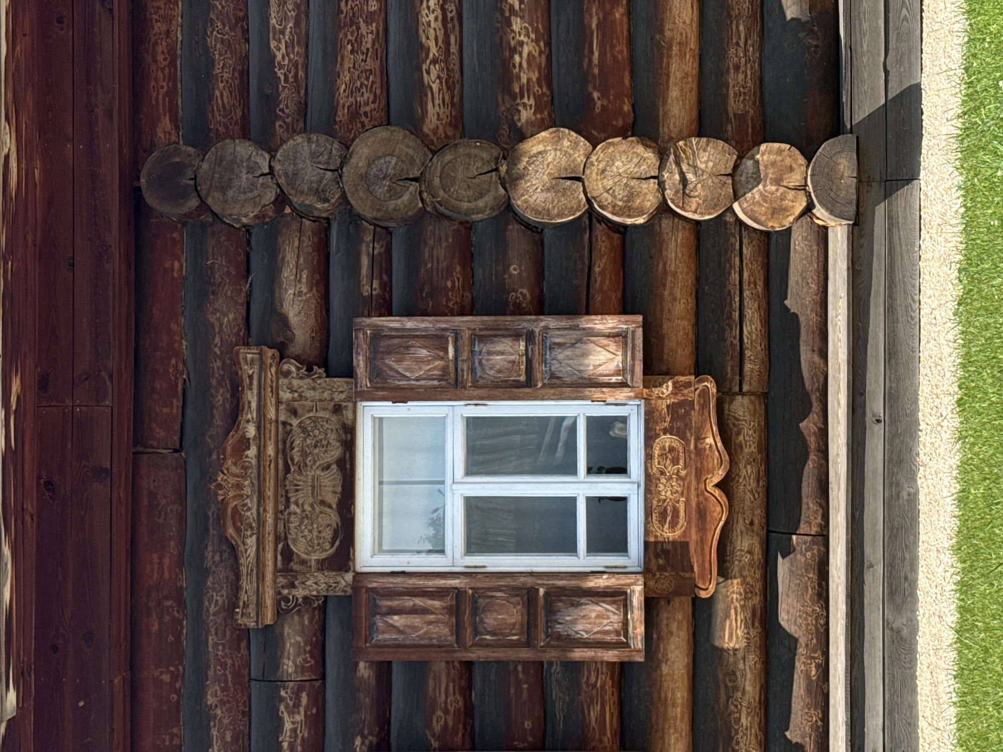 This is a medium shot, eye-level, full-frame, horizontal photograph of a wooden window on the side of a log cabin. The cabin is made of dark, weathered logs. The window has white trim and white shutters that are partially open. The window frame is ornately carved with decorative patterns. To the right of the window, there is a vertical stack of round, wooden logs. In front of the cabin, there is a wooden porch with a gravel pathway and a patch of green grass. The image is well-lit with natural sunlight, casting shadows on the wooden porch and the front of the cabin. The scene appears to be in a rural or historical setting. This is an image of the Shushenskoye Museum-Reserve in Shushenskoye, Krasnoyarsk Krai, Russia, a place associated with the exile of Vladimir Lenin.