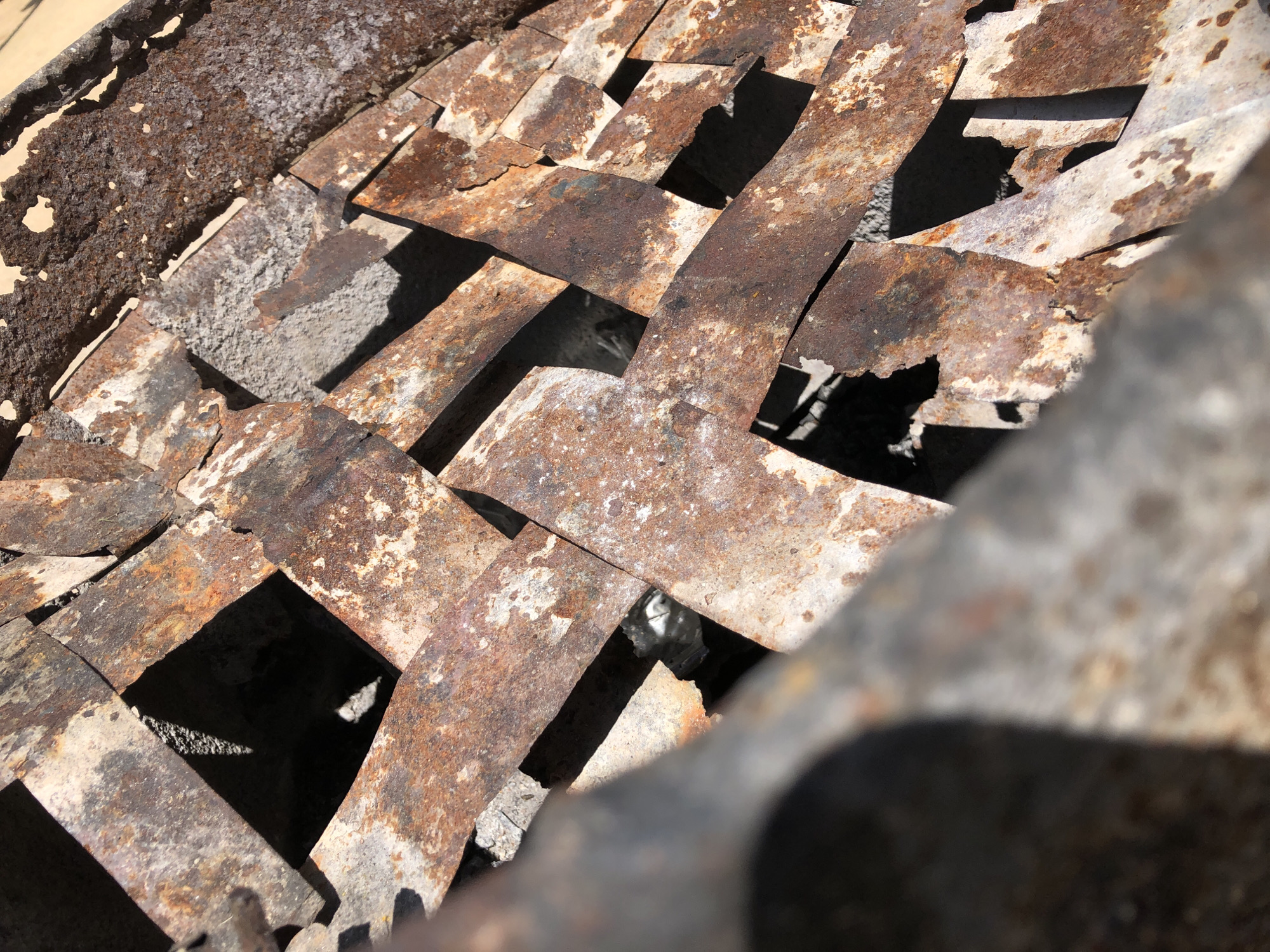 This is a close-up, high-angle, outdoor photograph of a woven metal structure, likely a grate or screen, showing significant rust and decay. The metal strips are overlapping, creating a pattern of square and rectangular openings through which shadows and darker areas are visible. The texture of the rusted metal is rough and pitted, with patches of lighter, possibly original, metal or primer showing through. The lighting suggests it is daytime, casting strong shadows. There is no visible indication of the city of Jos, Nigeria, nor are there any people, actions, or interactions depicted. The scene is purely an abstract composition of decaying metal.