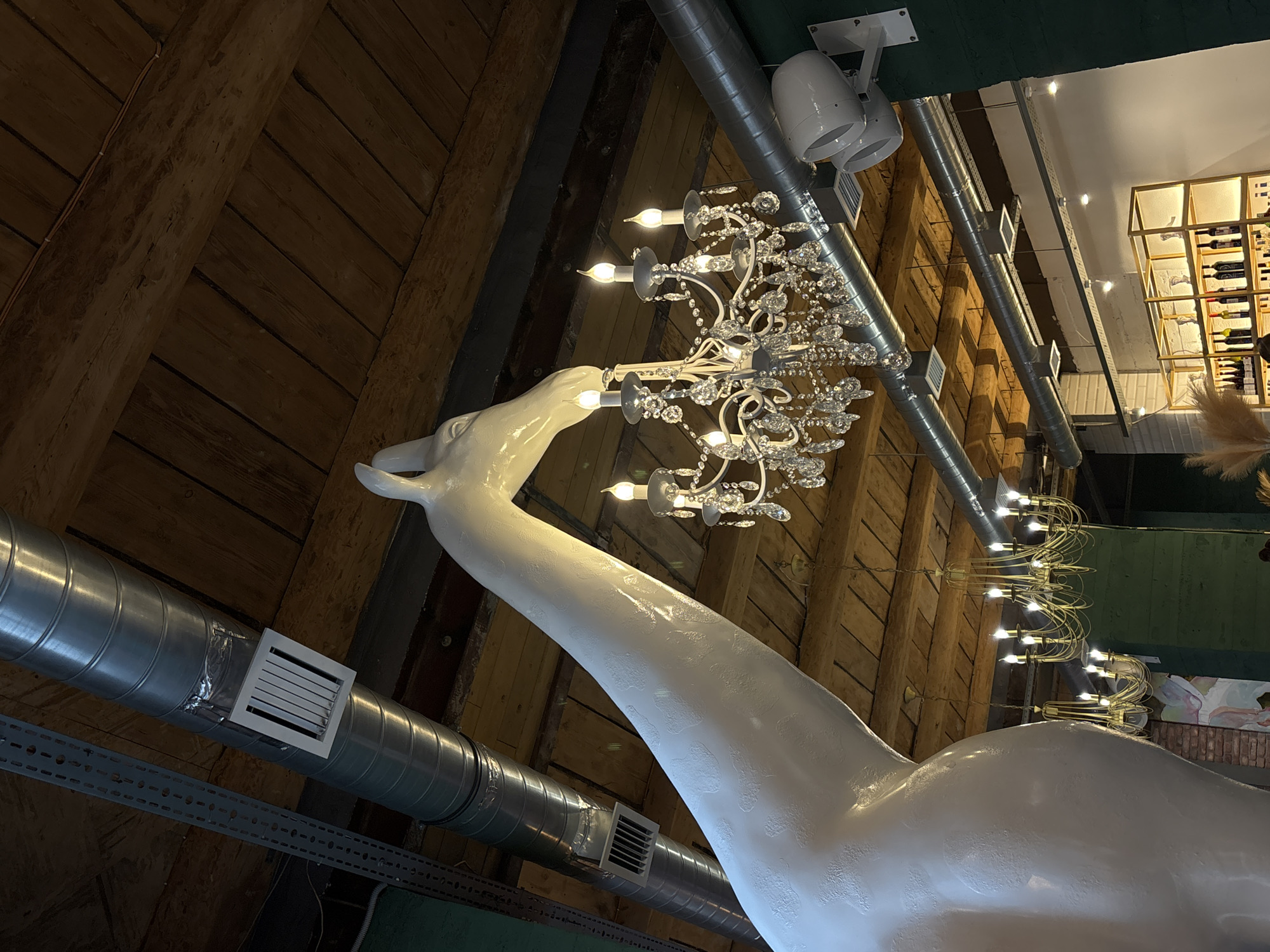 A low-angle shot reveals an ornate chandelier adorned with numerous crystal pendants and faux candle lights, mounted atop a large, glossy white sculpture of a swan's head and neck. The swan sculpture curves upward against a background of rustic wooden planking and industrial metal ventilation ducts. Above the chandelier, the ceiling transitions to a modern interior with track lighting and a shelving unit displaying wine bottles. The setting appears to be a stylish, unconventional space, likely a restaurant or bar. The location is the restaurant "Swan Princess", in Saint Petersburg, Russia.