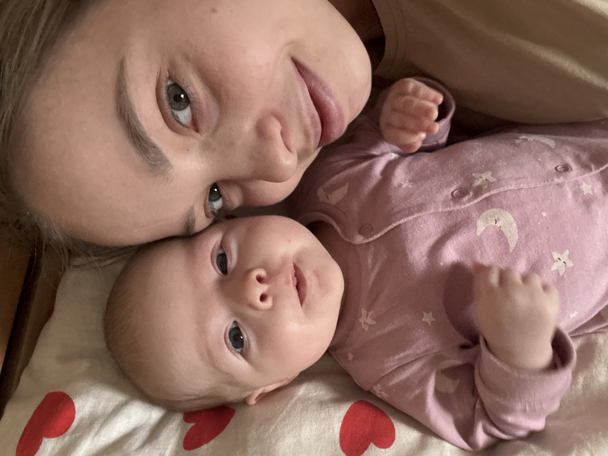 A close-up, tilted shot shows an adult and a baby lying on a white blanket with red heart patterns. The adult, fair-skinned, with grey eyes and a light complexion, has their head close to the baby. The baby, also fair-skinned with grey eyes, is wearing a pink onesie decorated with white stars and crescent moons. The baby is lying on its back with its hands in fists. The photo appears to be taken indoors, possibly in a home. The location is unspecified but claimed to be in the city of Chaikovsky, Russia.