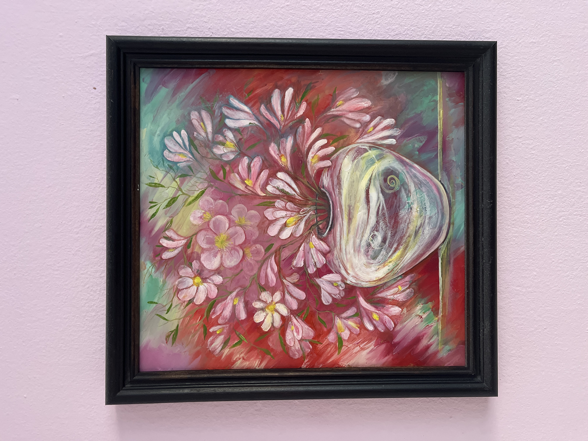 A framed painting of flowers in a vase hangs on a pink wall. The painting depicts pink and white flowers arranged around a bulbous, transparent vase. The background is a mix of red, green, and purple brushstrokes. The frame is black. The location is Burgas, Bulgaria.