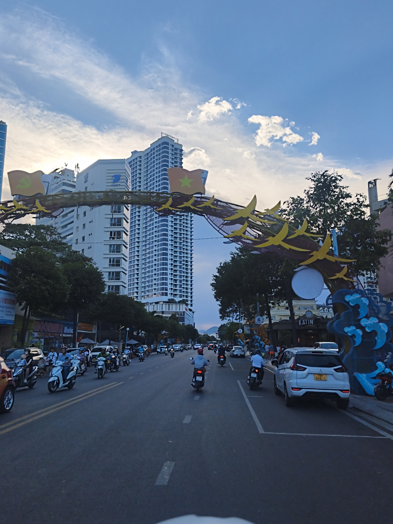 This is a street-level, outdoor, vertical, full shot captured during daylight with a bright sky and some clouds. The scene depicts a busy street in Nha Trang Ward, Vietnam, possibly during a celebration or event, indicated by an arched decorative structure spanning the road, adorned with flags and yellow bird-like ornaments. A line of motorcycles and cars is moving along the asphalt road, with pedestrians visible on the sidewalks. Tall buildings, including a prominent white high-rise with blue accents, line the street. The overall impression is one of urban activity and festivity.