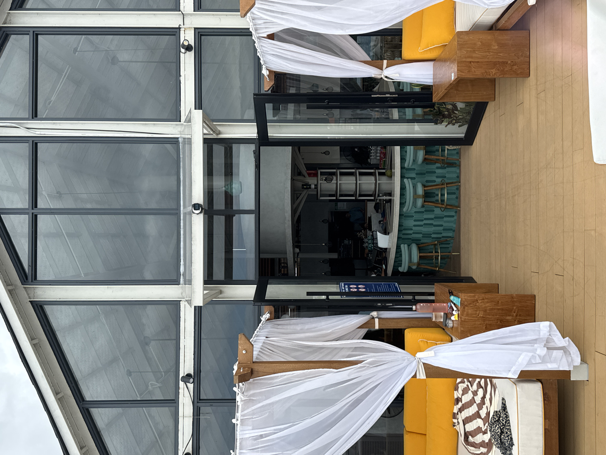 The image captures an outdoor seating area with cabanas at a resort in North Kuta, Indonesia. The scene features two cabanas with white sheer curtains draped over wooden posts, framing views of a building with large glass windows. In the foreground, a wooden deck leads to the cabanas, each furnished with a yellow cushioned lounge and a wooden side table. On the side table, there is a pink water bottle, a glass, and some tubes of what appear to be toiletries. Inside the building, a bar with teal tiled walls and stool seating is visible through open double doors. The overall ambiance suggests a relaxed, tropical setting.