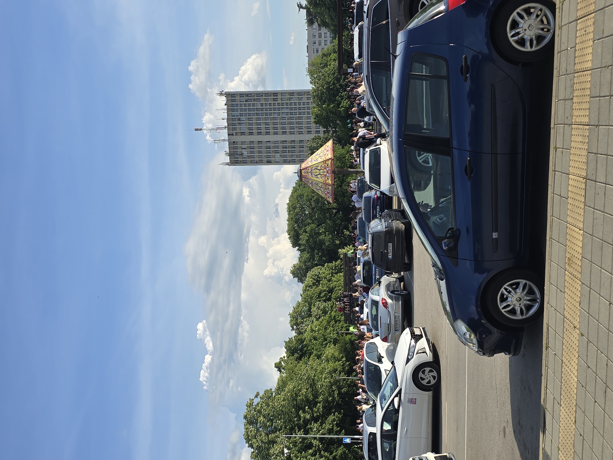 The image shows a parking lot filled with cars on a sunny day. A crowd of people is gathered beyond a line of trees in the background. A tall, gray building with antennas is also visible, along with an ornate, decorative structure. The photograph appears to have been taken in an urban environment.