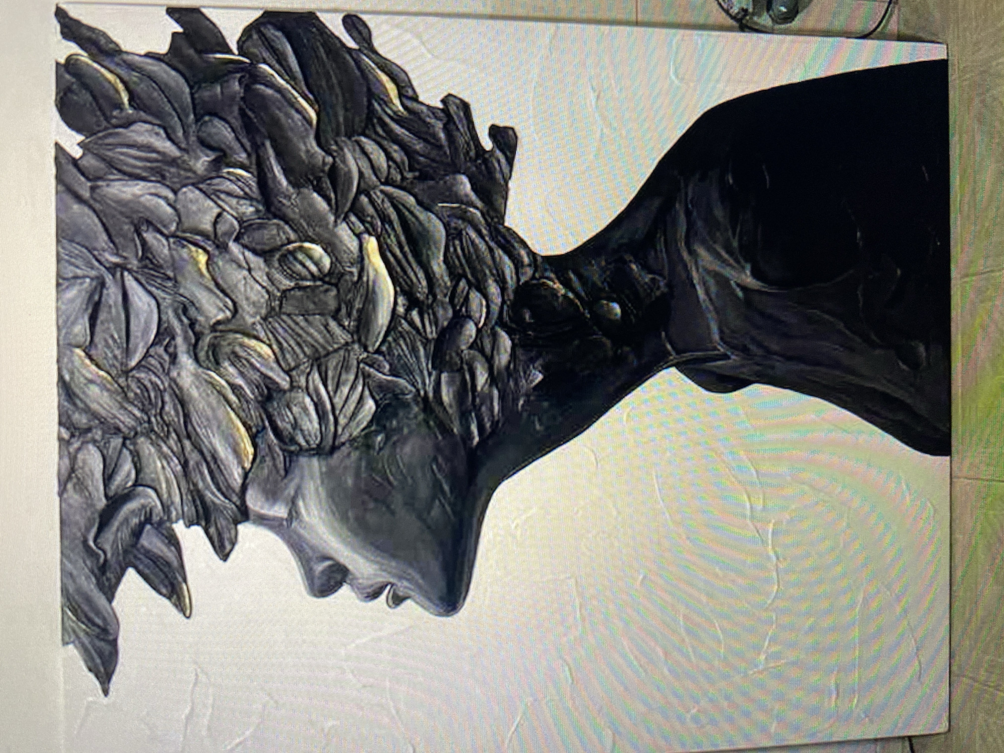 A close-up, side-profile, full-shot photograph shows a black and gray textured artwork of a woman's head and upper torso. The artwork is set against a white background with a subtle textured pattern. The woman's face is smooth and sculpted, with an open mouth and closed eyes. Her hair is depicted as a mass of layered, dark gray, textured elements that extend upwards and outwards from her head, with highlights of a metallic gold color. The neck and shoulders of the woman are rendered in a solid black, matte finish. The overall texture of the artwork suggests a sculpted or relief application of material. The background has horizontal, wavy lines of texture. The artwork is displayed on a surface that appears to be a floor or a stand, with hints of a wall behind it. The lighting appears to be overhead, casting shadows that enhance the three-dimensional quality of the artwork.