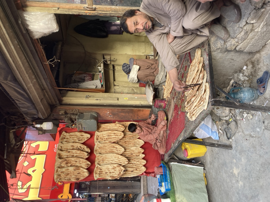 The image depicts a street scene in Quetta City Tehsil, Pakistan, centered on an open-fronted flatbread (naan) stall. A man in a light brown shalwar kameez squats in the foreground on the right, looking directly at the camera while arranging baked flatbreads on a low table covered with a red rug, using a pair of tongs. To his left, a young boy, also wearing a light brown shalwar kameez, sits cross-legged on the same table. Behind them, numerous baked flatbreads are displayed vertically in rows against a red fabric panel on the shop's exterior wall, beneath a red sign with Urdu script. A gas meter is affixed to the wall adjacent to the bread display. Inside the dimly lit shop entrance, an individual partially obscured by traditional clothing is seated. The ground surface is paved but uneven and shows debris, including a yellow plastic jerry can, a blue and white sack, a blue ceramic pitcher, and blue sandals. A small green side table holds a white bottle and a blue plastic container.