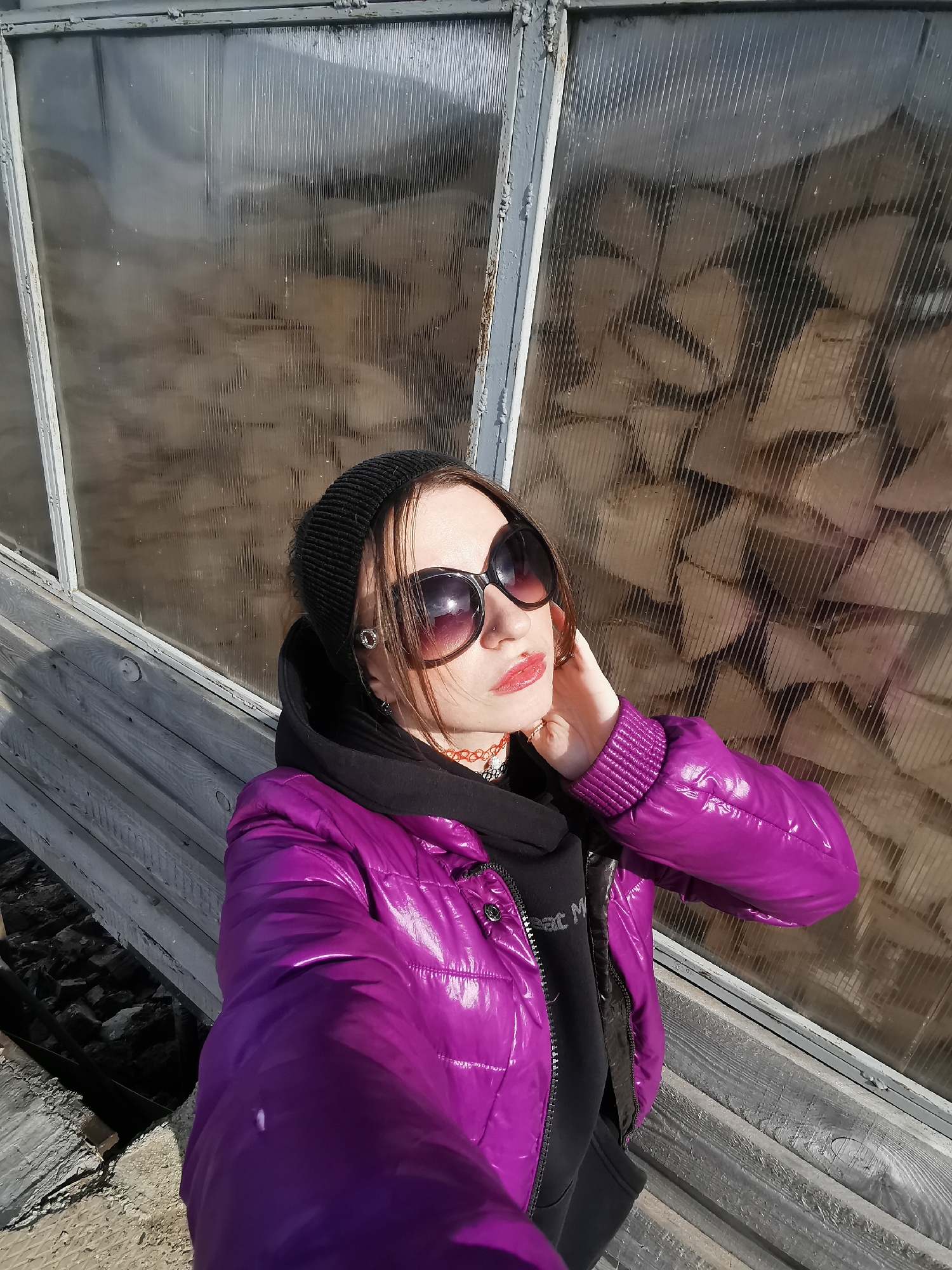 A person is posing for a photo in front of a glass window that is obscuring a stack of firewood. The person is wearing a black beanie, large sunglasses, a black hoodie, and a shiny purple puffer jacket. Their right hand is raised towards their neck. The photo appears to be taken outdoors, with weathered wooden planks visible below the window. The location and city are unknown.