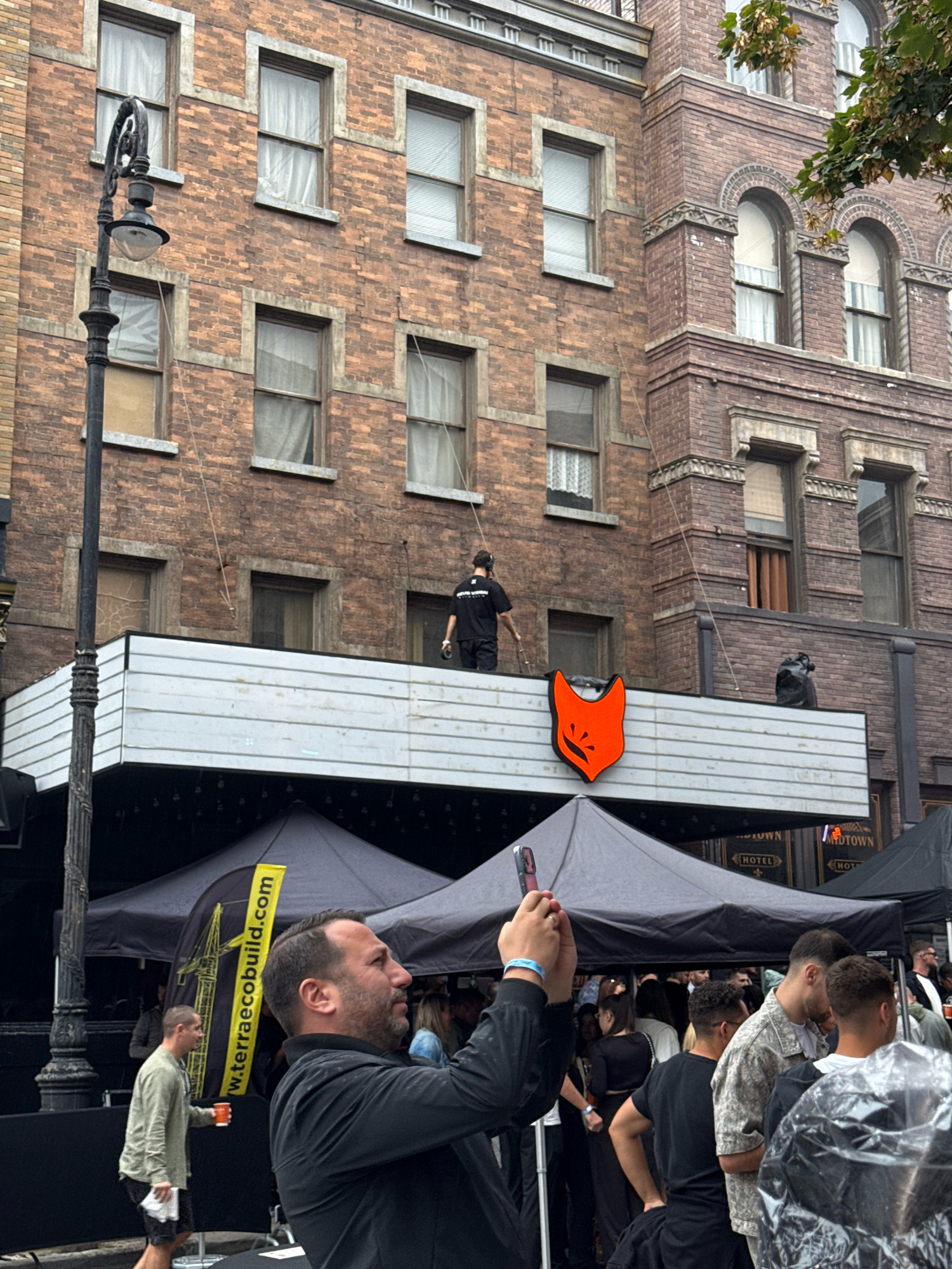 In Sofia, Bulgaria, an outdoor scene unfolds with a brick building as the backdrop. A man in a black shirt and shorts is visible on the roof of a white structure, holding onto wires. Below him, an orange fox logo is displayed. Several tents are set up on the ground level. In the foreground, a man in a black shirt is taking a photo with his phone. Other people are gathered under the tents, some in focus and others out of focus. A lamppost stands to the left of the frame. The overall impression is one of an organized event taking place in an urban setting.