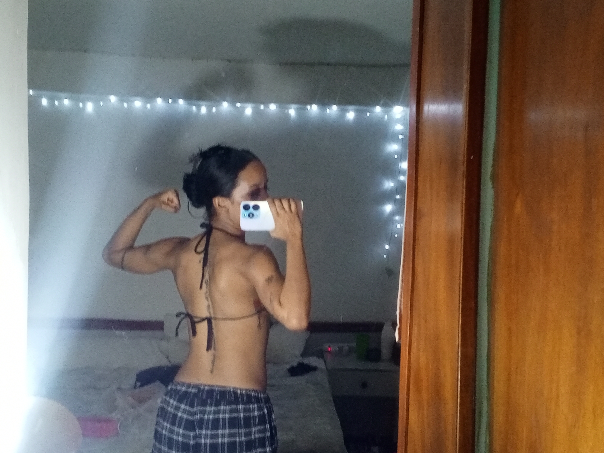 A woman is reflected in a mirror. She is wearing a bikini top and plaid pajama bottoms. She is holding a smartphone in front of her face and posing, flexing her arm. In the background are string lights, a bed, and a bedside table. The image appears to be taken indoors. There is no evidence to indicate that the scene is in Porlamar, Venezuela.