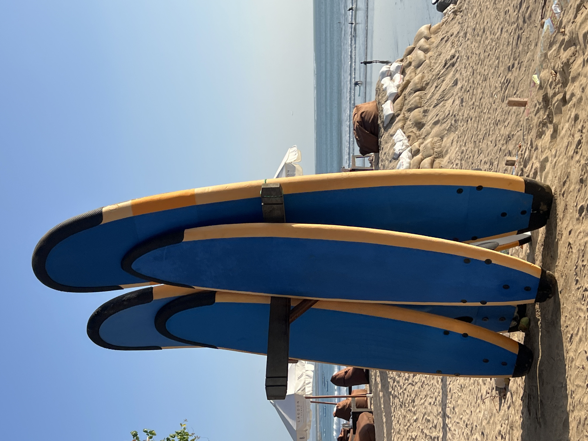 The image captures a sunny day on Kuta Beach, Indonesia, with a clear blue sky above. In the foreground, several blue surfboards with yellow accents are propped up on a sandy dune, held by a dark wooden rack. The surfboards are oriented vertically, with their fins facing the sand.
The beach is populated by a few individuals engaged in various activities. In the middle ground, several people are visible in the ocean, some appearing to be surfing or wading. Further back, a white umbrella shades a seating area with brown beanbag chairs and a table, suggesting a relaxed beachside establishment. The ocean stretches to the horizon, with gentle waves lapping at the shore. Sandbags are also present on the dune, possibly for coastal protection. The overall scene depicts a typical day at a popular Indonesian beach destination.
