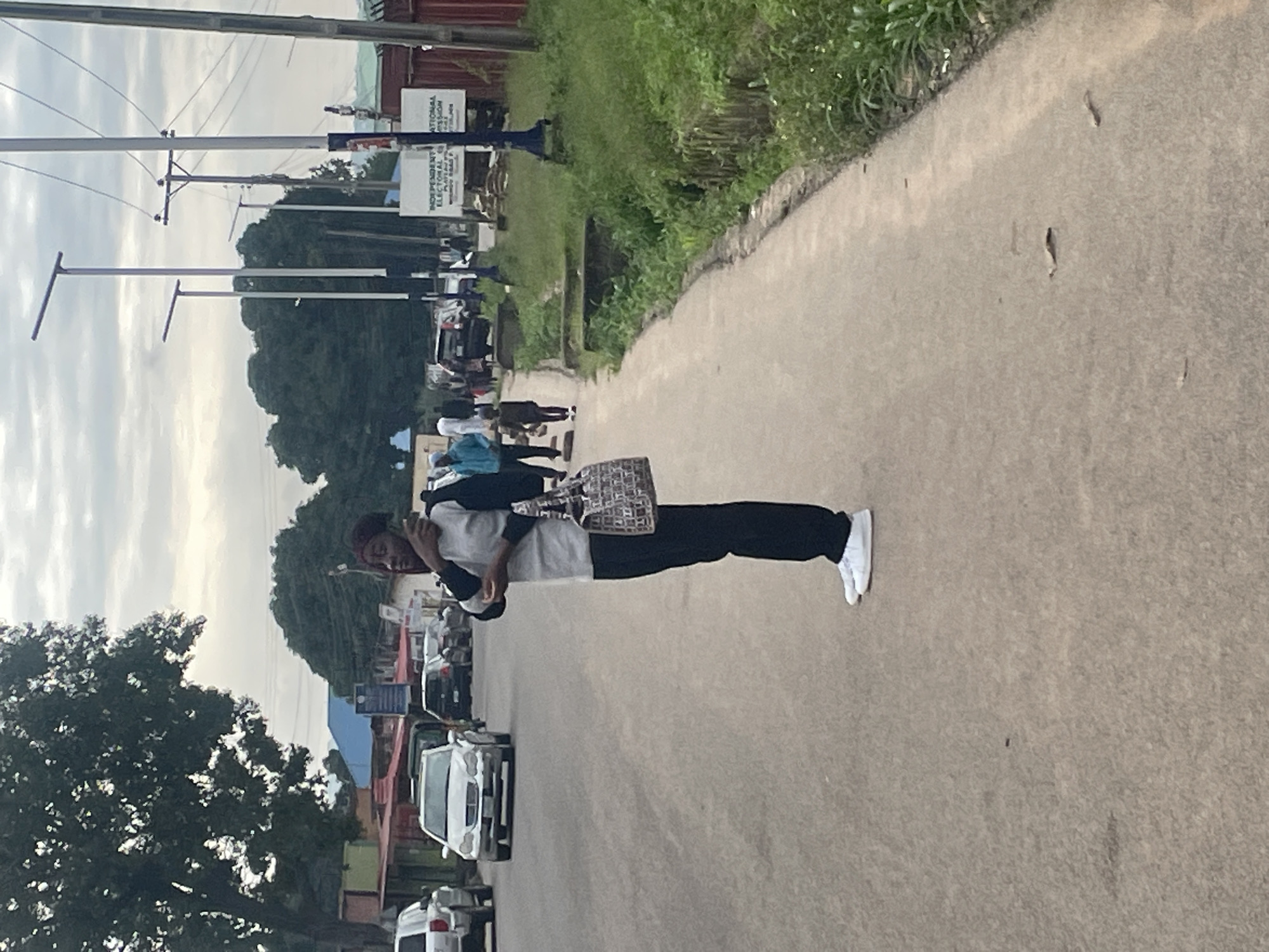 A young Black person stands on a paved road in Jos, Nigeria, on a cloudy day. They are looking to their left while holding a dark-colored cellphone to their ear. They wear a grey long-sleeved shirt with dark sleeves, dark pants, and white sneakers. A patterned tote bag hangs from their left shoulder. The road is lined with grassy verges and a few streetlights. Several cars are parked and in motion on the road, and other pedestrians are visible in the background. Signs with text are mounted on poles. The area appears to be a commercial or administrative district.