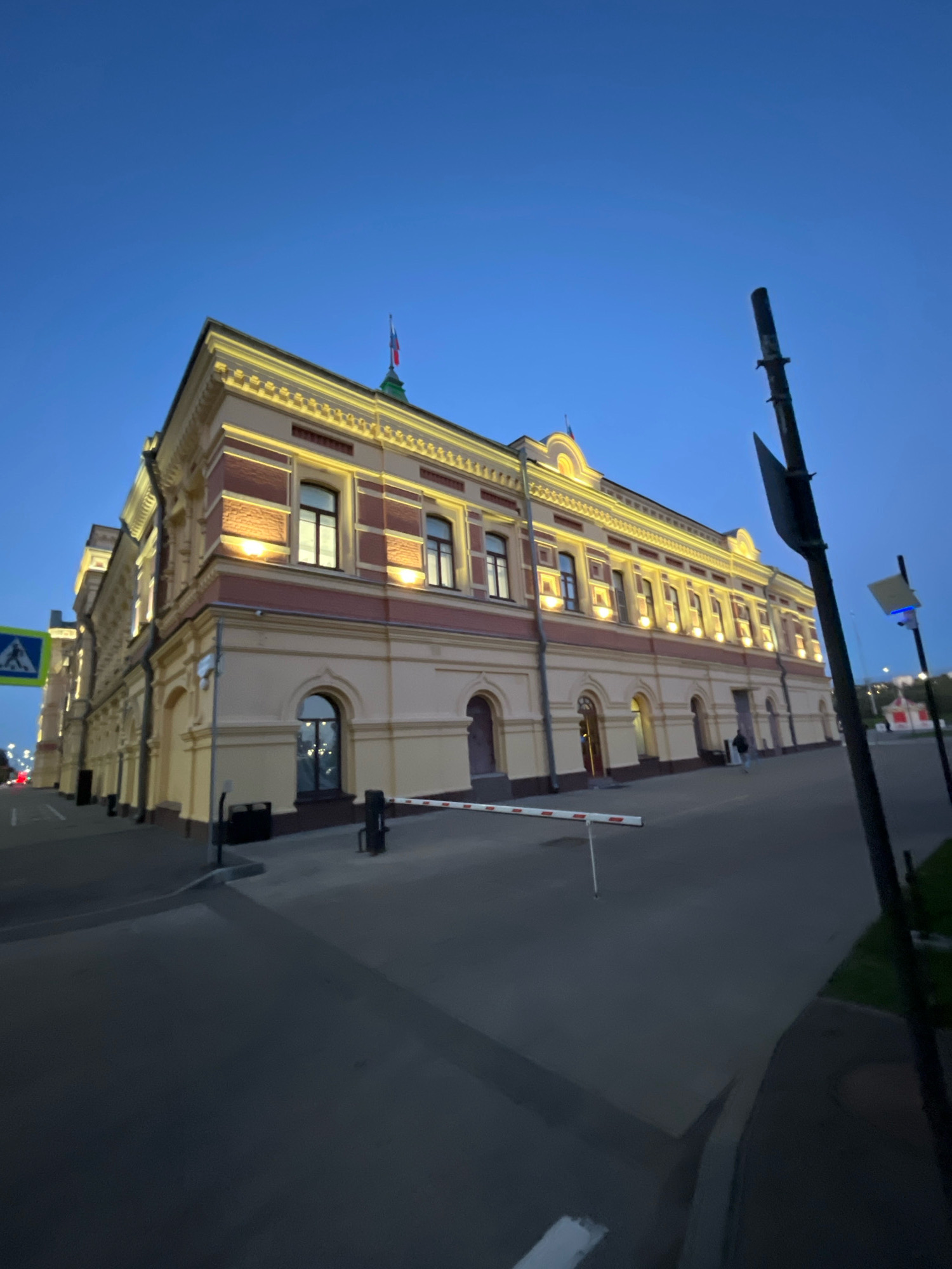 The image depicts the Nizhny Novgorod State Academic Drama Theater named after Maxim Gorky, illuminated by exterior lights against a twilight sky. The building, an imposing classical structure, features a light yellow facade with red accents and ornate architectural details along the roofline. Several windows are visible, some reflecting the internal lights. In the foreground, a paved area with a red and white striped barrier suggests a controlled entrance. A dark pole, possibly a lamppost or signpost, stands prominently on the right side of the frame, with a smaller, illuminated sign on a pole further down the street. A pedestrian walkway is visible to the left, marked with a blue and white pedestrian crossing sign. The overall impression is one of a well-maintained, historic building bathed in the warm glow of artificial lighting as evening descends.