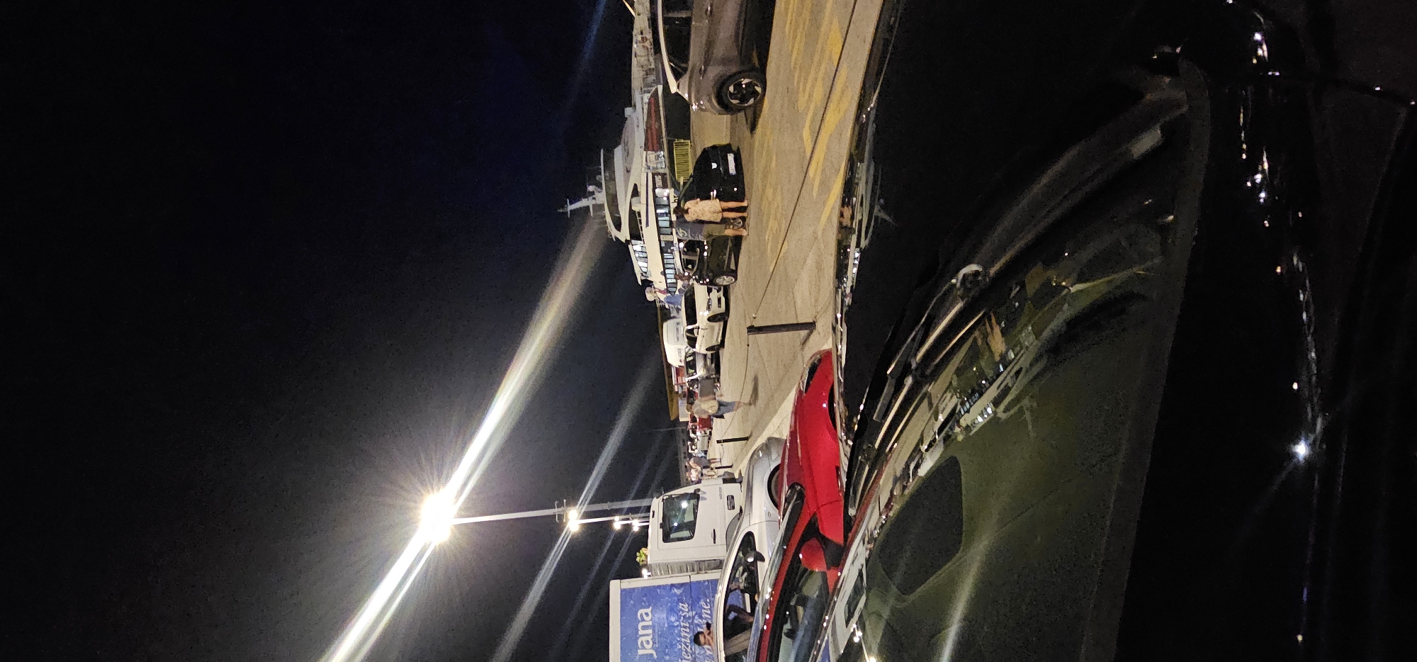 The image depicts a nighttime scene at a ferry port in Split, Croatia. In the foreground, the hood of a black car is visible, reflecting the surrounding lights. Behind it, a red car is parked, followed by a line of other vehicles including white and black cars, and a white delivery truck. The truck has the logo "Jana" on its side.
Further back, a large white ferry is docked. Several people are walking and standing near the vehicles and the ferry. The scene is illuminated by bright streetlights that cast long beams across the pavement. The sky is dark, indicating it is nighttime. The overall impression is of a busy port with vehicles waiting, possibly to board the ferry.