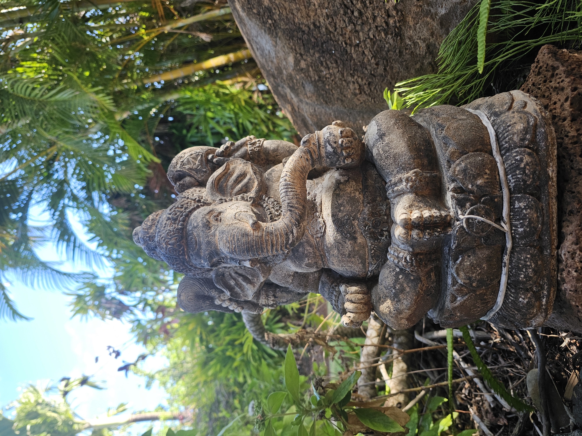 A stone statue of Ganesha, a deity with an elephant head, sits in a cross-legged position on a lotus-shaped base in a garden. The statue is weathered and appears to be made of dark, rough stone. Ganesha is depicted with multiple arms, some holding objects. The statue is surrounded by lush green foliage, palm trees, and bamboo stalks, suggesting a tropical or subtropical environment. A large, rough-textured rock is visible to the right of the statue. The sky is visible in the background, with some sunlight filtering through the leaves. The scene is captured in an outdoor setting, likely a garden in Napili, United States of America.