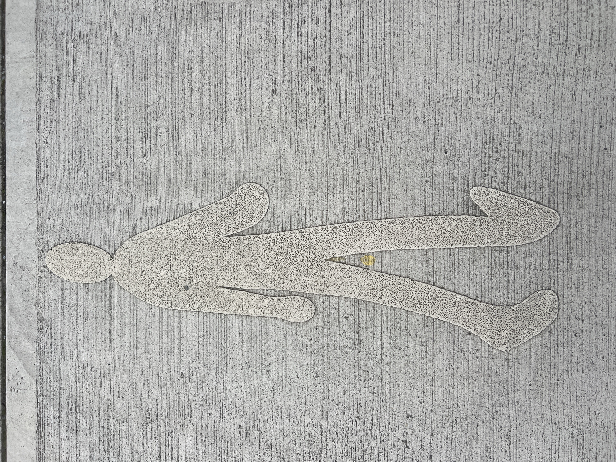 A schematic representation of a walking person is imprinted on a textured, grey concrete surface. The figure is outlined in a lighter shade of grey, with a stippled texture within the outline. The figure faces towards the right of the frame, with its left leg extended forward and its right leg back. The left arm is bent at the elbow and extends forward, while the right arm is bent and angled towards the body. The surface it is imprinted on appears to be a sidewalk or pedestrian pathway. The image is captured from a top-down perspective, showing the texture of the concrete with its horizontal striations. There is no indication of the specific location within Balbriggan, Ireland.