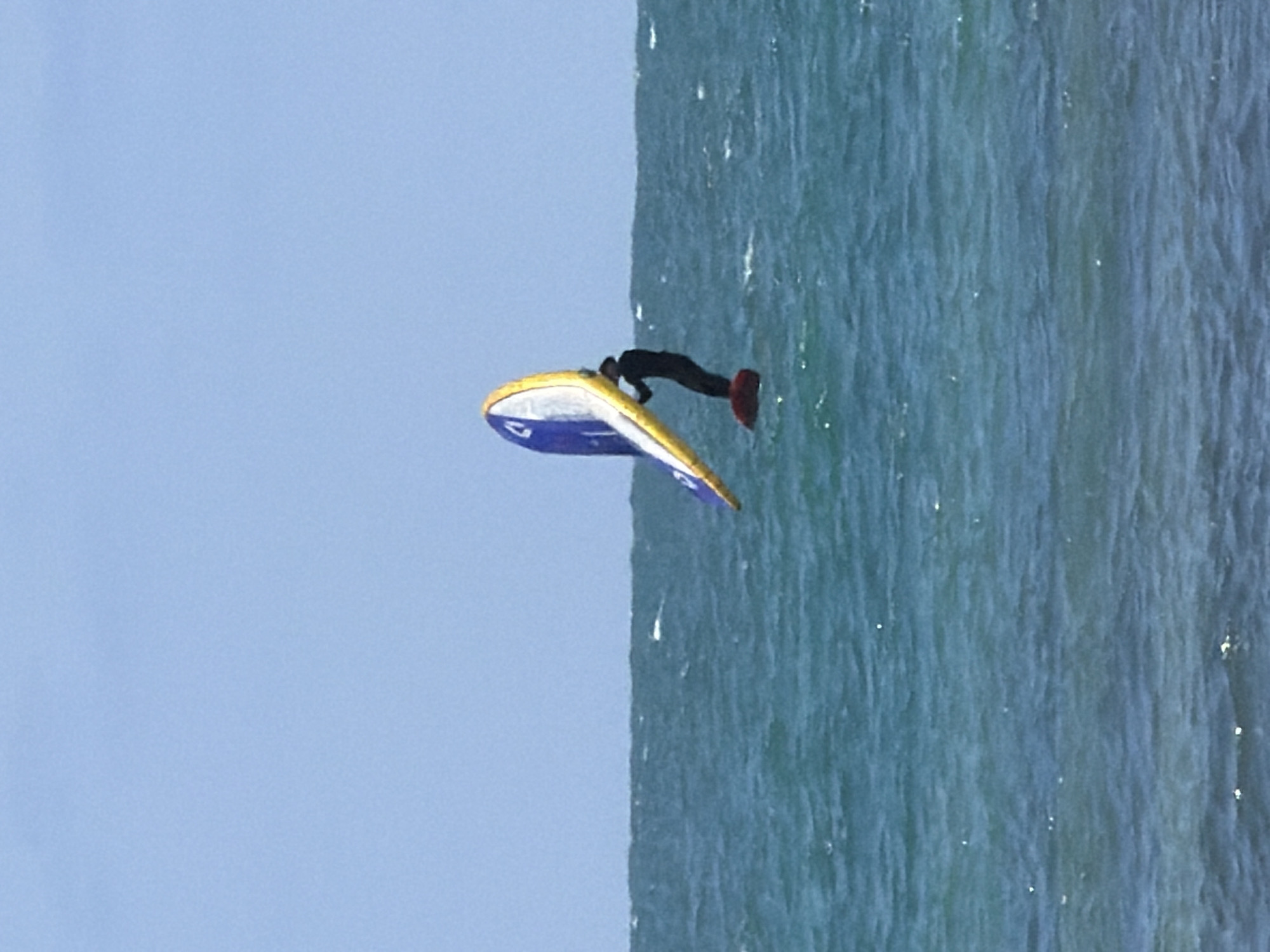 A person in a black wetsuit and red booties is seen lying on a yellow and blue hydrofoil board on the water. The location is most likely Tel-Aviv, Israel, as it is a coastal city. The water is a turquoise blue. There is a clear, light blue sky. The image appears to be taken from a high vantage point.