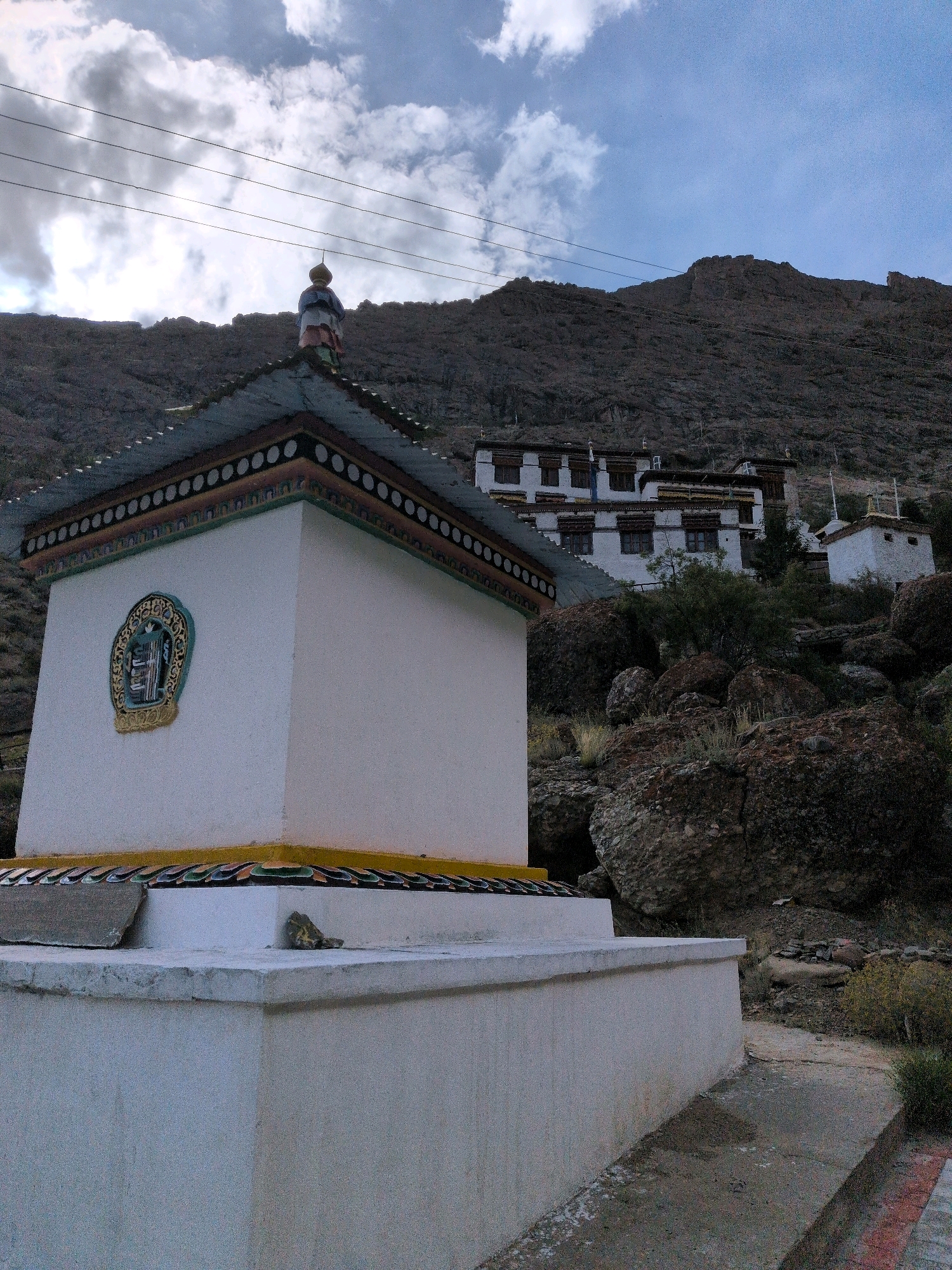 An eye-level, medium outdoor shot captures a white stupa in the foreground. The stupa has a decorative roof with a yellow trim and a patterned band of circles. On the side of the stupa, there is a round, ornate emblem. Behind the stupa, a rocky hillside rises, dotted with sparse vegetation. In the middle ground, a monastery complex with white buildings and dark-framed windows is nestled into the hillside. A figure stands on the roof of a small structure in the far background. The sky above is cloudy and grey. The scene appears to be in Tanglakpurna, India.