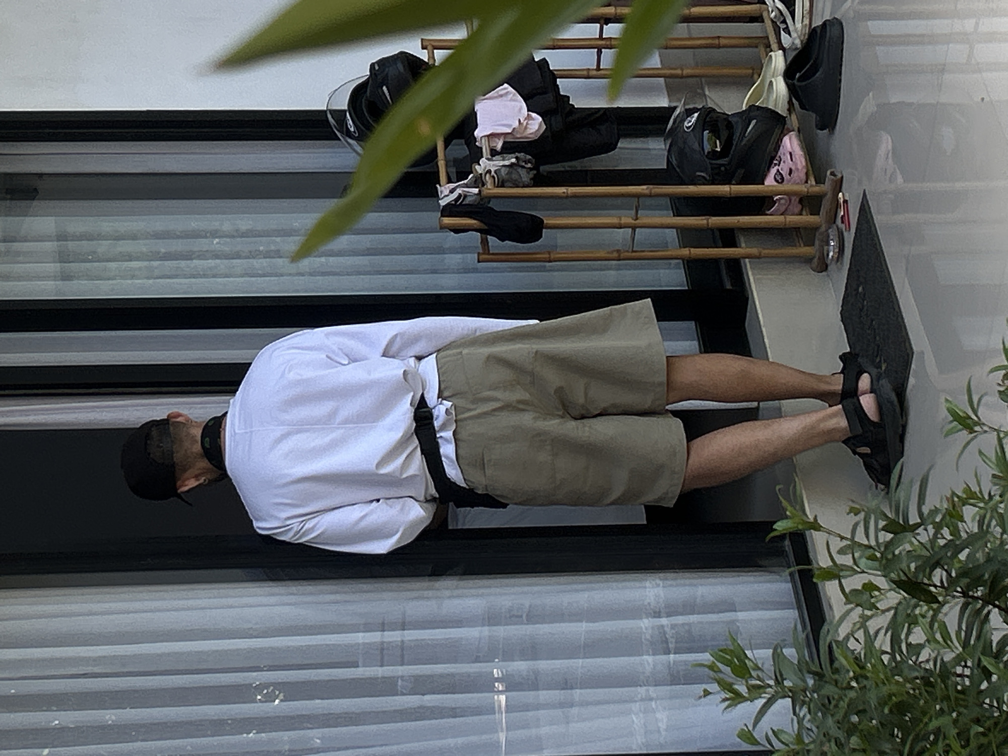 A person stands facing a white, vertical-slatted door. They are wearing a white long-sleeved shirt, khaki shorts, a black fanny pack around their waist, a black baseball cap, and black sandals. Next to the door is a small wooden rack with several items of clothing and two helmets. In the foreground, there are green plants and a gray floor. The scene appears to be outdoors, possibly in front of a residence or a shop in North Kuta, Indonesia.
