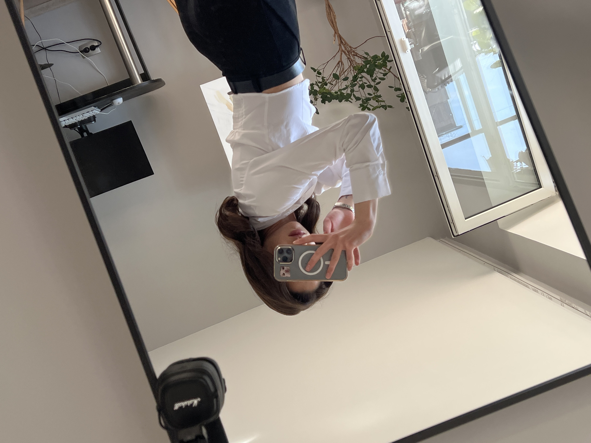The image depicts a woman in a white collared shirt and black pants taking a mirror selfie. She is holding a phone in front of her face, obscuring it. To her left, a large window with a white frame reflects a bright blue sky and greenery. A small, potted tree with sparse branches sits to the left of the woman. In the background, a black computer monitor is visible on a desk. Above the mirror, a pair of black headphones with "Marshall" branding hangs. The overall setting appears to be indoors, possibly a room in a modern apartment or studio. The location is not discernible from the visual information presented in the image and therefore cannot be identified as Ternopil, Ukraine.