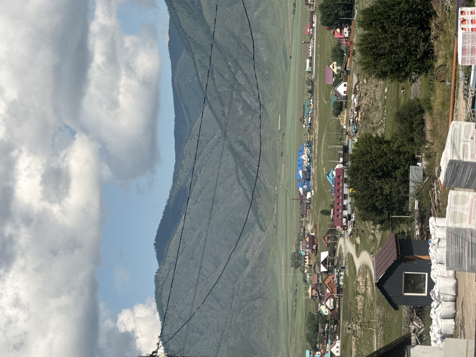 The image depicts a rural landscape near Onguday, Russia, featuring a village nestled against a backdrop of rolling, tree-covered mountains under a cloudy sky. In the foreground, a small, dark shed with a brown roof and a single window is visible. In front of the shed, stacks of white bags, likely containing construction materials, are arranged on the ground. Further into the scene, a winding dirt road leads through the village, which is characterized by a collection of houses with colorful roofs, ranging from blue and red to brown and white. Some houses appear to be under construction. The village is spread across a green, grassy plain that extends towards the mountains. In the mid-ground, two cows graze in a field. Utility lines are strung across the sky in the middle ground. The overall environment suggests a remote, possibly agricultural, settlement.