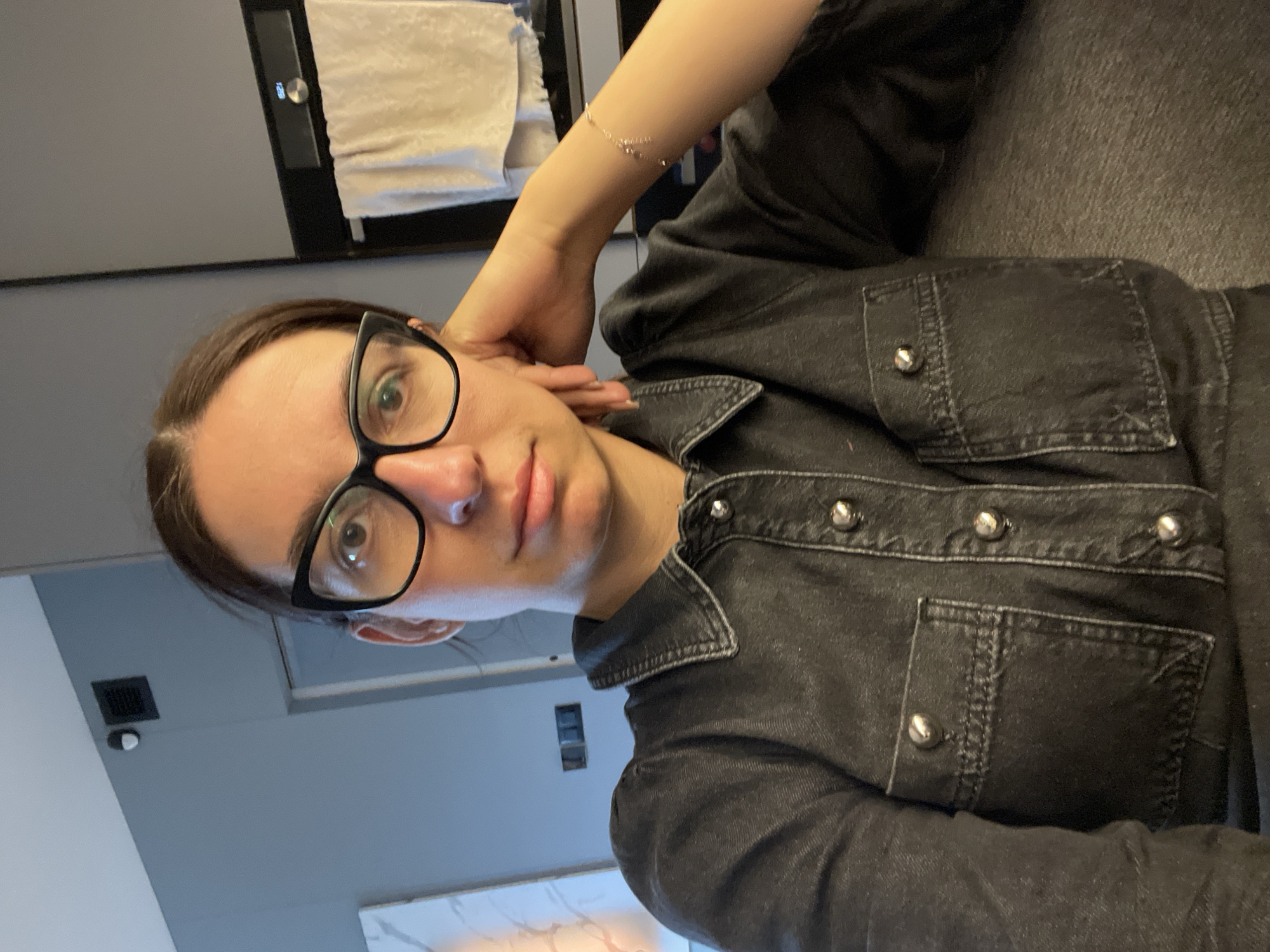 The image, rotated 90 degrees clockwise from a typical upright portrait, captures a person reclining indoors, looking directly at the camera. The subject is a young to middle-aged individual with dark hair, styled neatly away from the face, and wears prominent black-framed glasses. Their expression is neutral and direct. They are dressed in a dark, possibly black denim-style jacket or shirt, featuring several metallic silver buttons and visible chest pockets with buttoned flaps. A delicate, chain-link bracelet is worn on their right wrist, with the arm bent and the hand resting near their head.
The background indicates a modern indoor setting. To the left, a dark-colored appliance, resembling an oven or microwave, shows a digital display with the time "12:05". A white, textured fabric, possibly a towel, is partially visible above it. To the right, a dark grey, textured surface, possibly a sofa or upholstered wall, occupies a significant portion. Lower in the frame, a light grey wall features a black ventilation grate and a small white dome-shaped object, which could be a sensor or security camera. A subtle, light-colored surface with a marble-like pattern is visible at the bottom edge. The overall aesthetic is contemporary, utilizing a palette of greys, blacks, and whites. The setting is inferred to be in Sofia, Bulgaria, as provided.
