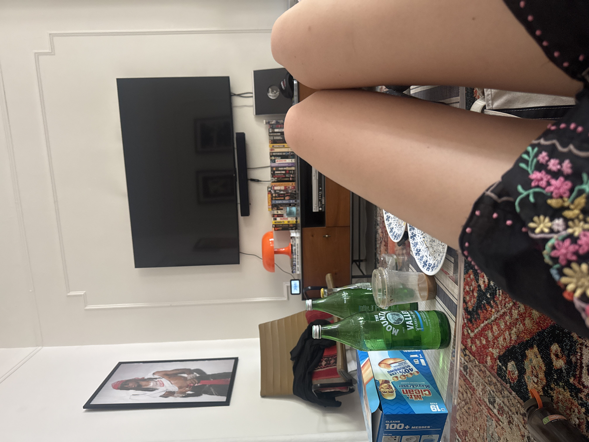 A person's legs are in the foreground, facing the camera. They are wearing black shorts with colorful floral embroidery. The person is sitting in a living room with a coffee table in front of them. On the coffee table, there are two bottles of Mountain Valley Sparkling Water, a plastic cup with a brown liquid, and two small plates with blue and white patterns. A box of Mr. Clean Magic Eraser is also on the table, along with a striped placemat. Behind the coffee table, there is a wooden media console with a TV mounted on the wall above it. The media console has several shelves with DVDs and a record player. An orange lamp is on the media console, and a framed picture of a person wearing a red bandana hangs on the wall to the left of the TV. A brown armchair with a black jacket draped over it is visible in the background. The scene is likely in New York City, United States of America.