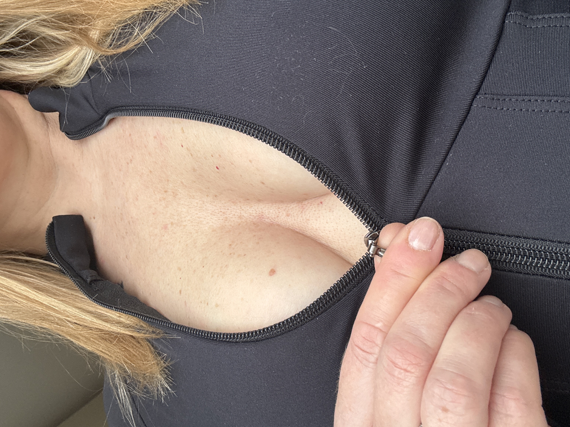 A close-up, slightly angled view shows a woman's partially exposed cleavage due to a partially unzipped black zippered garment. Her hand is visible, holding the zipper pull. A strand of blonde hair is visible on the left side of the image. The image does not provide any information that could be used to determine a location, specifically in Votkinsk, Russia.