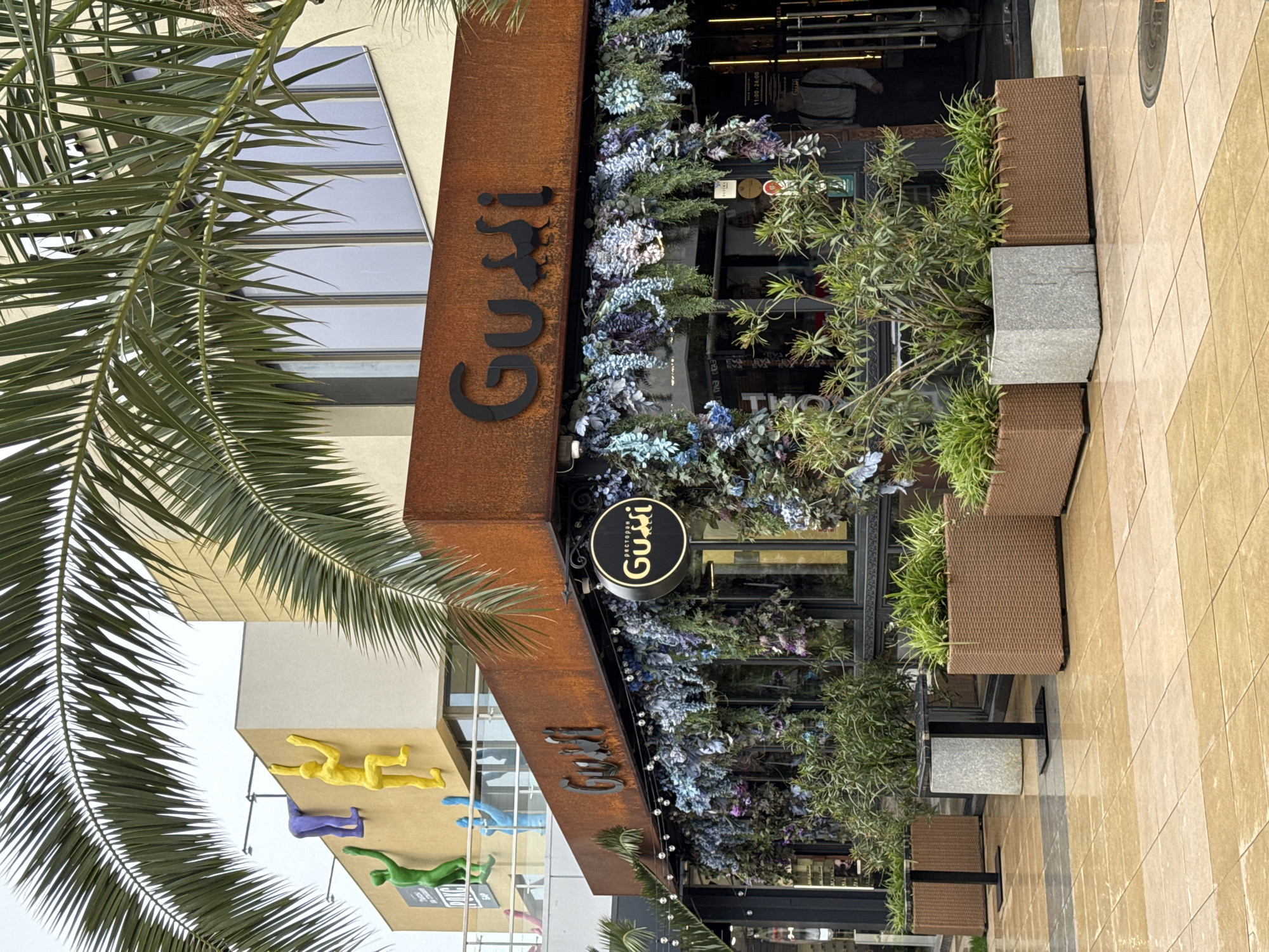 The image depicts the exterior of the "Gusi" restaurant in Sochi, Russia. The restaurant's facade is adorned with floral decorations and has a rusty brown signboard with the name "Gusi" in black lettering. A round sign with the restaurant's name in both English and Russian is also visible. Adjacent to the entrance are potted plants in brown rattan-style planters. The restaurant's windows reveal interior details. A palm tree partially obscures the left side of the frame. In the background, a building with colorful climbing figures is seen. The ground is paved with light brown tiles.