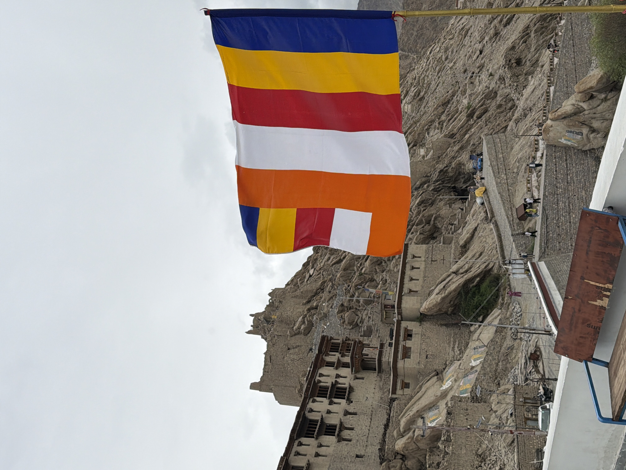 The image captures a view of the Thiksey Monastery complex, located in Thiksey, India. A large, colorful Buddhist prayer flag, with vertical stripes of blue, yellow, red, white, and orange, dominates the foreground, partially obscuring the rocky, mountainous landscape and the monastery buildings. The monastery itself is built into the hillside, with multiple levels of stone structures, featuring dark-paned windows and wooden balconies. A more ruined structure is visible higher up on the peak of the mountain. The sky is overcast and a muted grey. Several people are scattered across pathways and near buildings within the monastery complex, suggesting ongoing activity. A white car is parked near the lower section of the complex. In the immediate foreground, a wooden bench with a blue metal frame is partially visible. The overall scene is one of a rugged, historical religious site under a cloudy sky, with the vibrant prayer flag adding a splash of color.