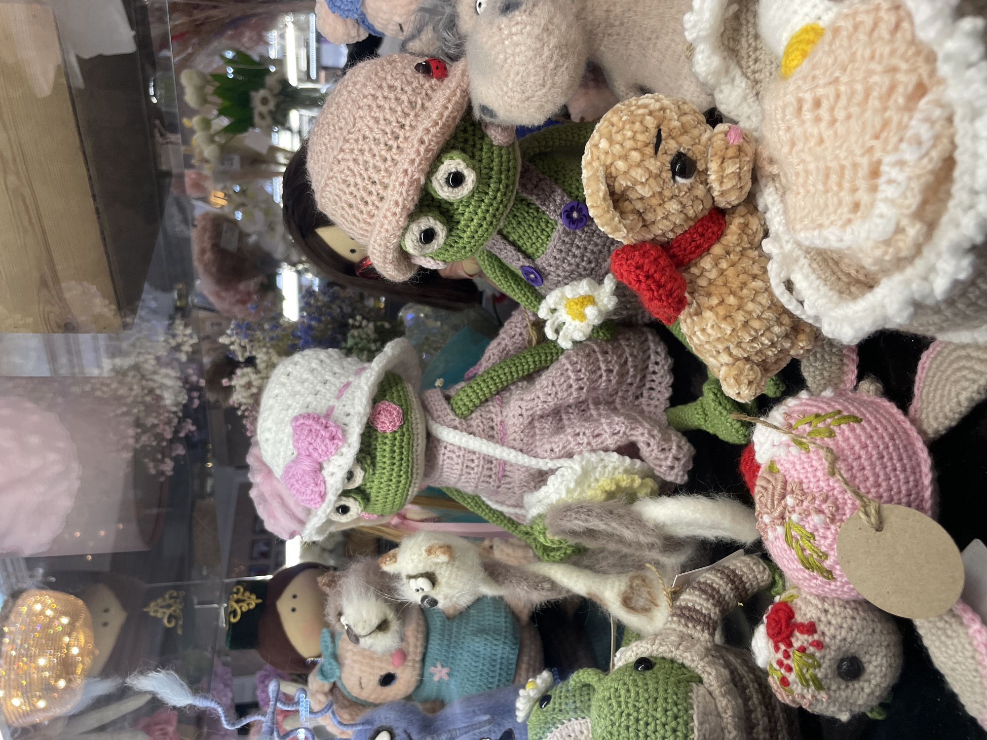 The image displays a close-up view of a collection of handcrafted crochet toys, arranged in a shop display. The toys include several frogs, a teddy bear, a cat, and other animals. The frogs are dressed in small hats and outfits, one with a pink dress and white apron, and another in purple overalls. The teddy bear is wearing a red scarf. The toys are situated on a dark surface, with some decorative elements and other shop merchandise visible in the blurred background, including what appears to be floral arrangements and lighting fixtures. The scene is well-lit, emphasizing the details and textures of the crochet work. The location is identified as Kazan, Russia.