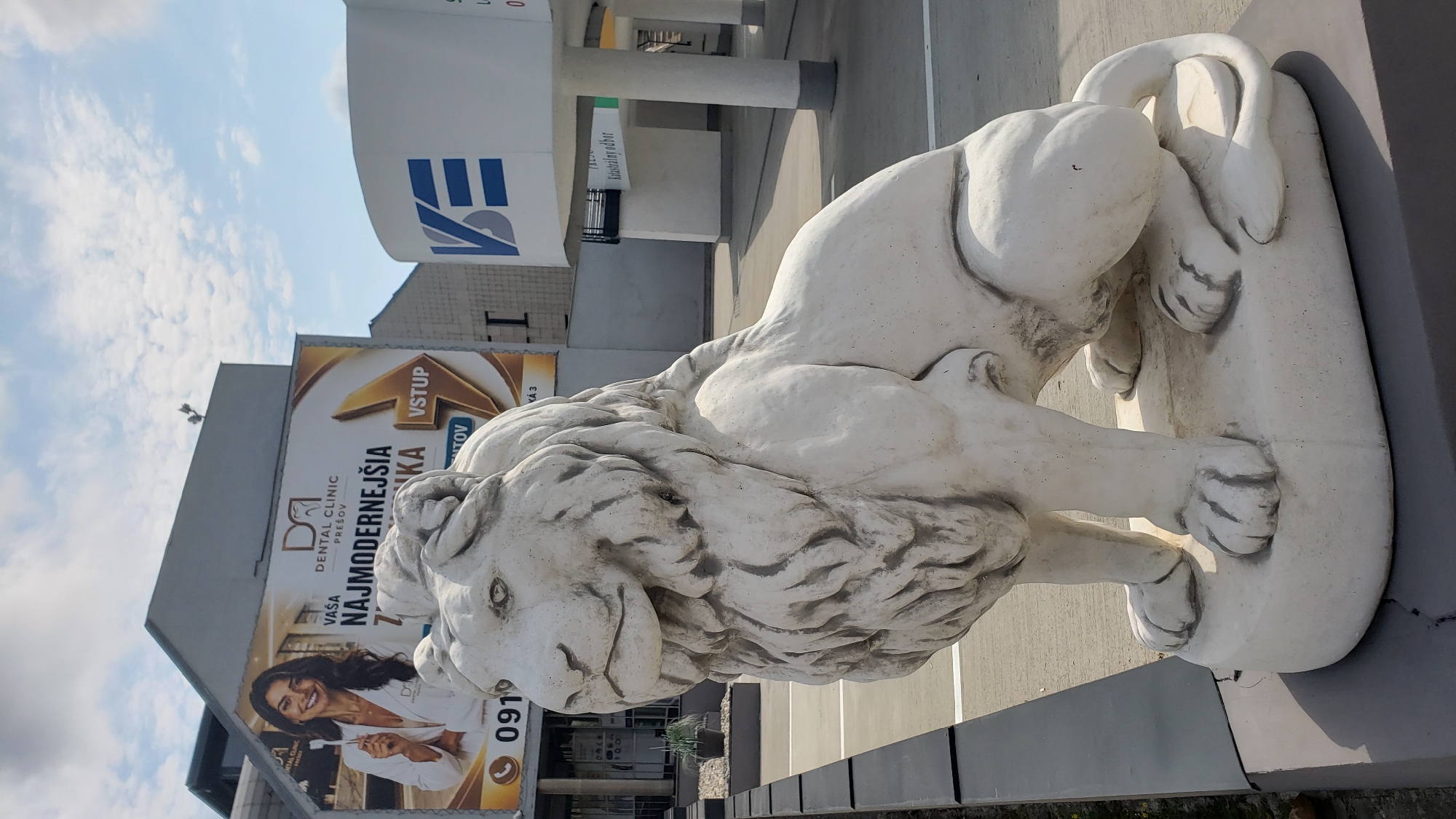 A white, concrete statue of a seated lion is positioned in the foreground, facing left. The lion has a prominent mane, detailed paws, and a tail curled around its rear. Behind the statue, a large billboard advertises "DENTAL CLINIC PREŠOV" with an image of a smiling woman holding a toothbrush. To the right of the billboard is a modern building with a large, white, curved structure displaying a blue logo that reads "VE". The scene appears to be an outdoor urban setting, possibly a parking lot or public space, under a bright, cloudy sky.