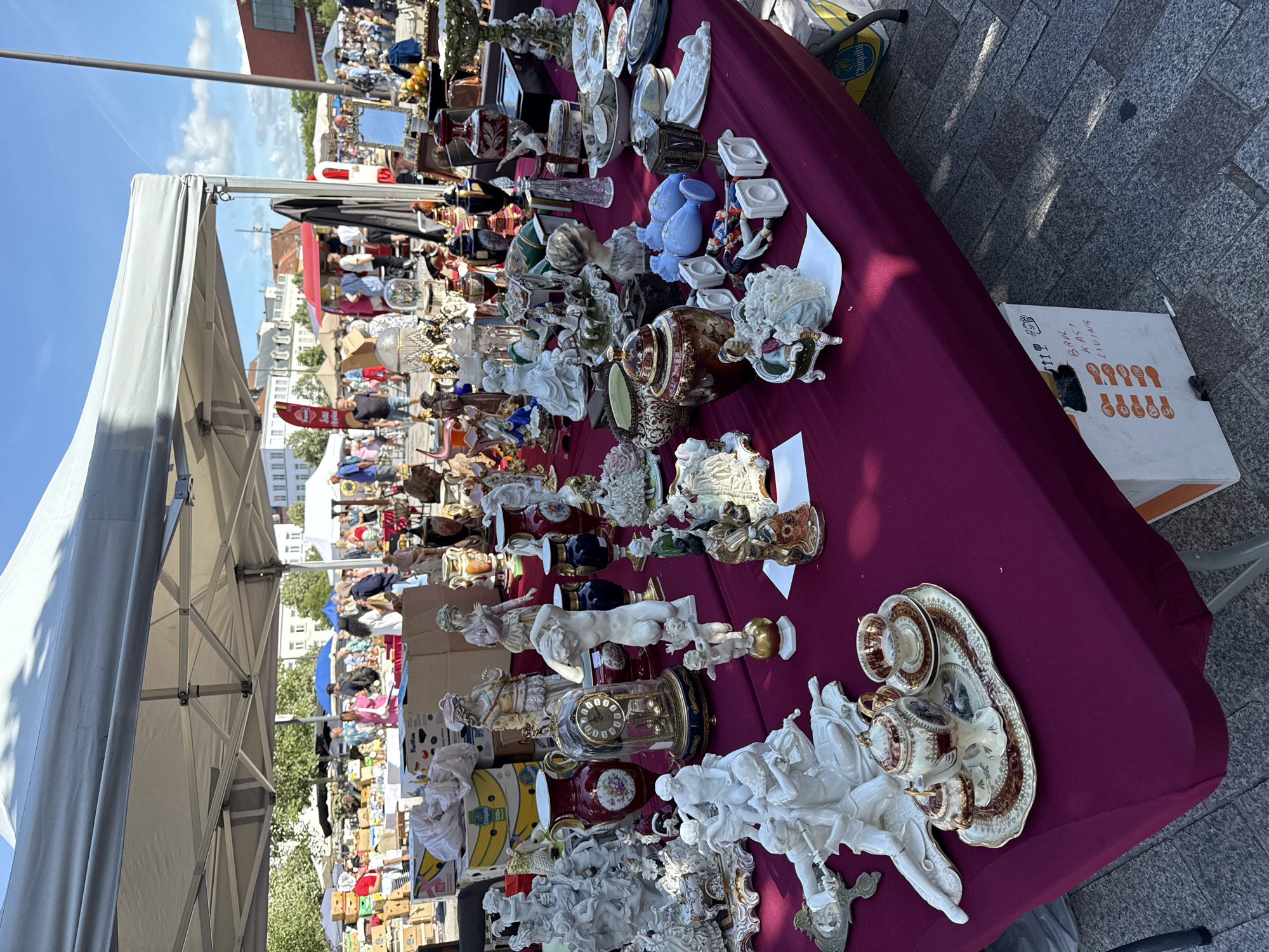 An outdoor market is displayed under a white tent. The foreground is dominated by a table covered with a deep red tablecloth, laden with a diverse assortment of porcelain figurines, vases, and decorative objects. Among these are several white porcelain sculptures of cherubs and people in classical poses, a detailed clock with a glass dome, ornate vases in red and blue, and a small tea set on a tray. The background shows other market stalls with various goods, and a crowd of people browsing. Buildings with traditional European architecture are visible in the distance, suggesting an urban setting. The ground is paved with stone.