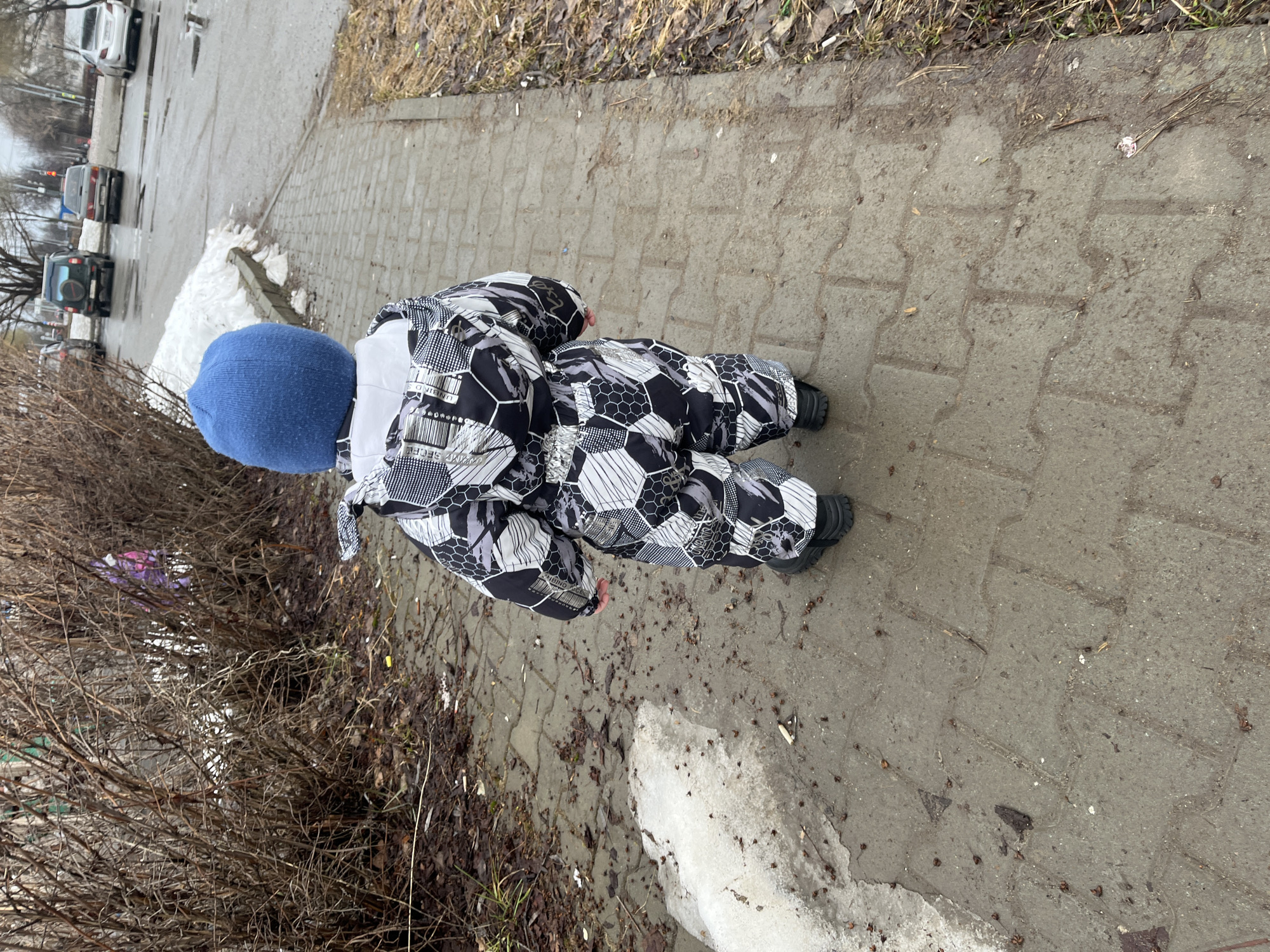 A child in a blue hat and black and white patterned snowsuit is standing on a sidewalk in Chaikovsky, Russia. The sidewalk is made of gray concrete pavers and has patches of melting snow along the edges. Adjacent to the sidewalk is a bare bush. In the background, cars are visible on a wet road. The weather appears to be overcast.